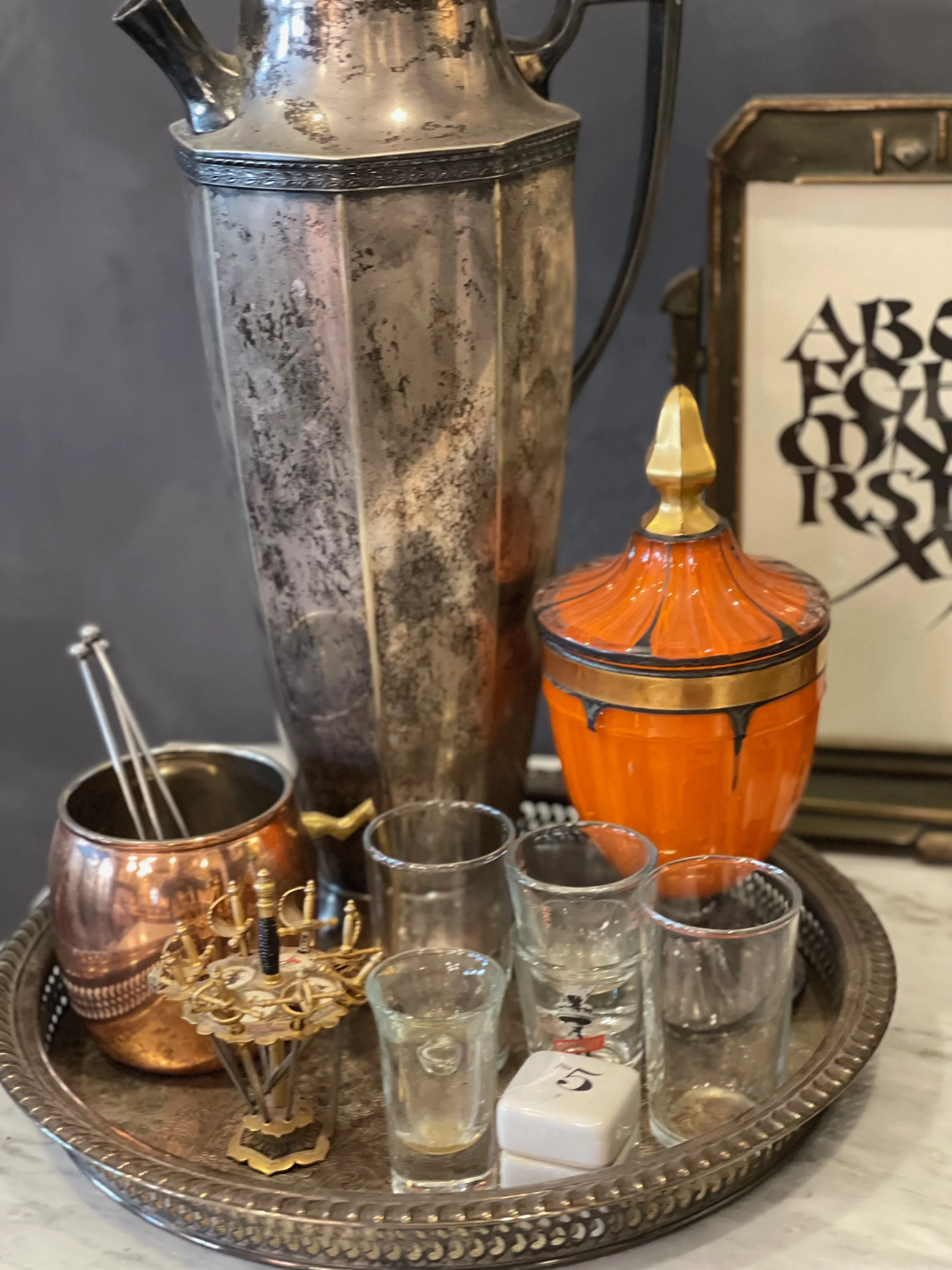Decorative tray with vintage items including a large tarnished metal beverage pitcher, an orange ceramic container with a lid, a copper cup, several small glasses, a small white cube, and a brass holder with utensils, all on a silver tray.