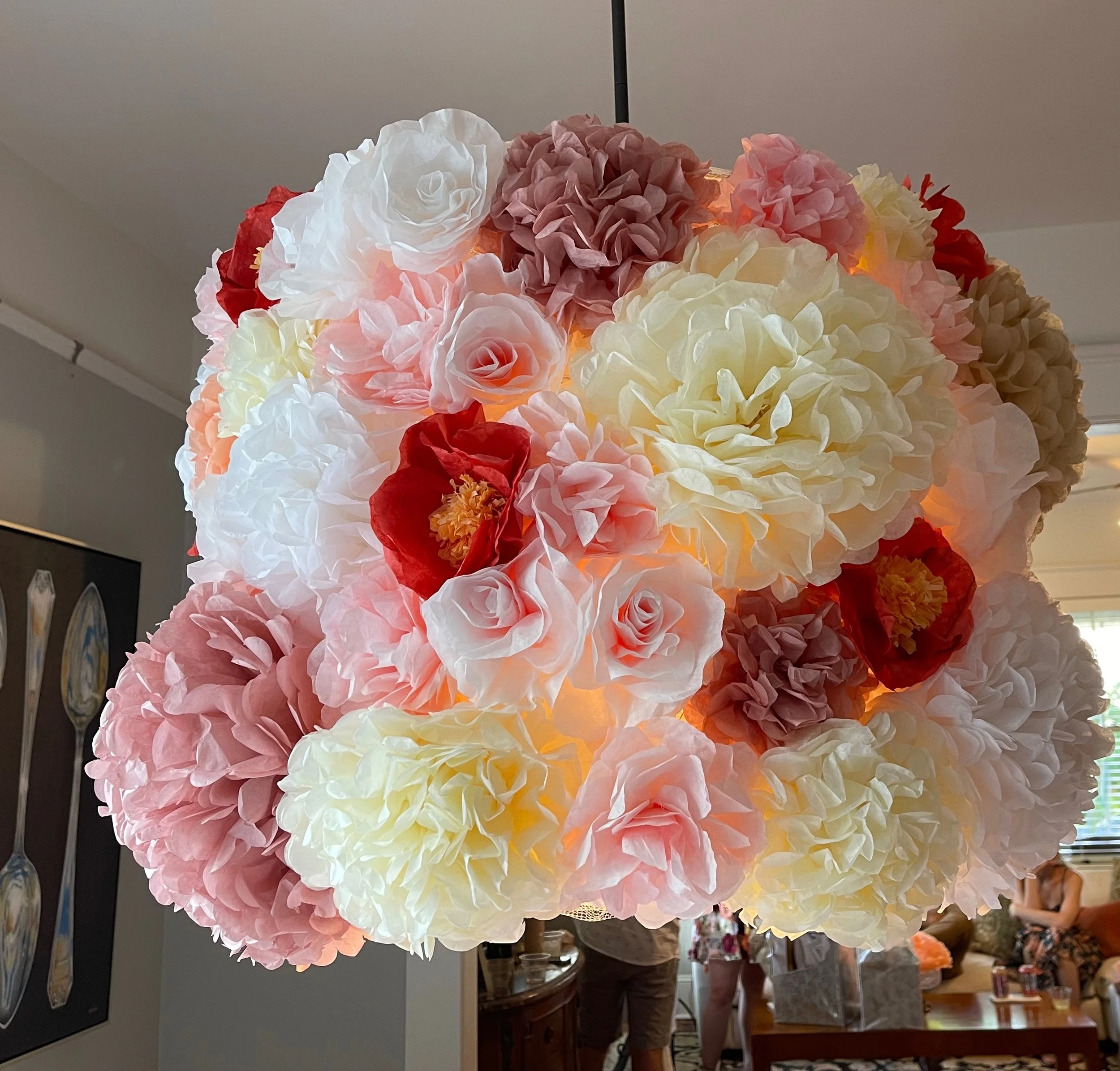 A spherical lamp made of pink, white, cream, red, and mauve paper flowers hanging in a room with people and decorations in the background.