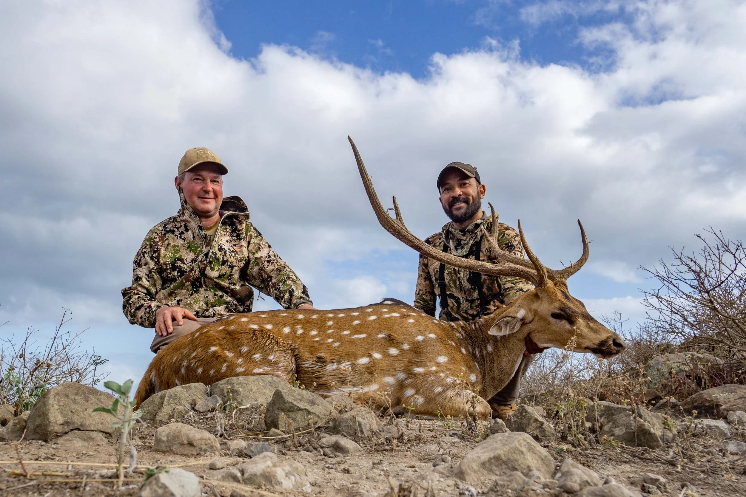 Two hunters pose behind an axis deer buck on a rocky Molokai ridge