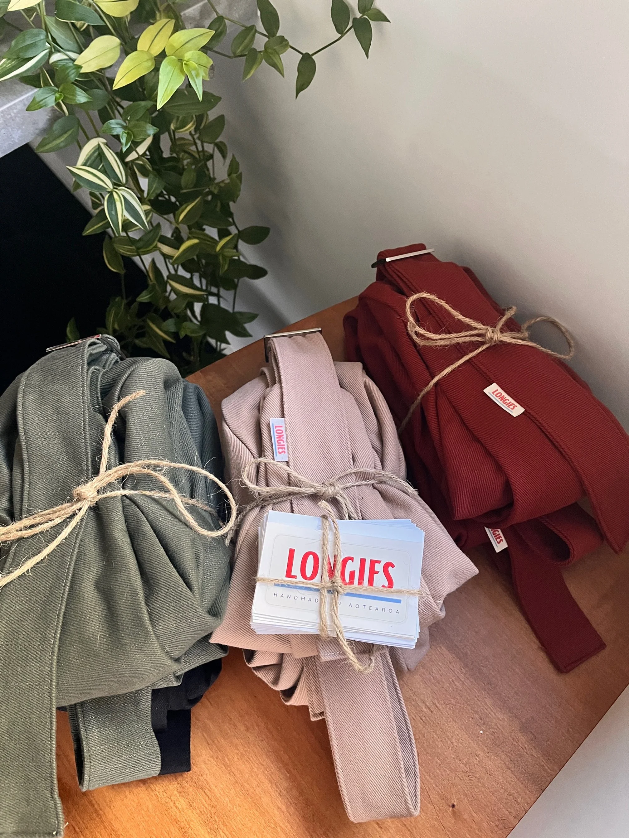 Three handmade denim Longies bags, ready to be sent to customers in olive green, dune sand, and ember red, each tied with twine, placed on a wooden surface near some green plant leaves. The pink pouch has a tag that says "LONGIES."