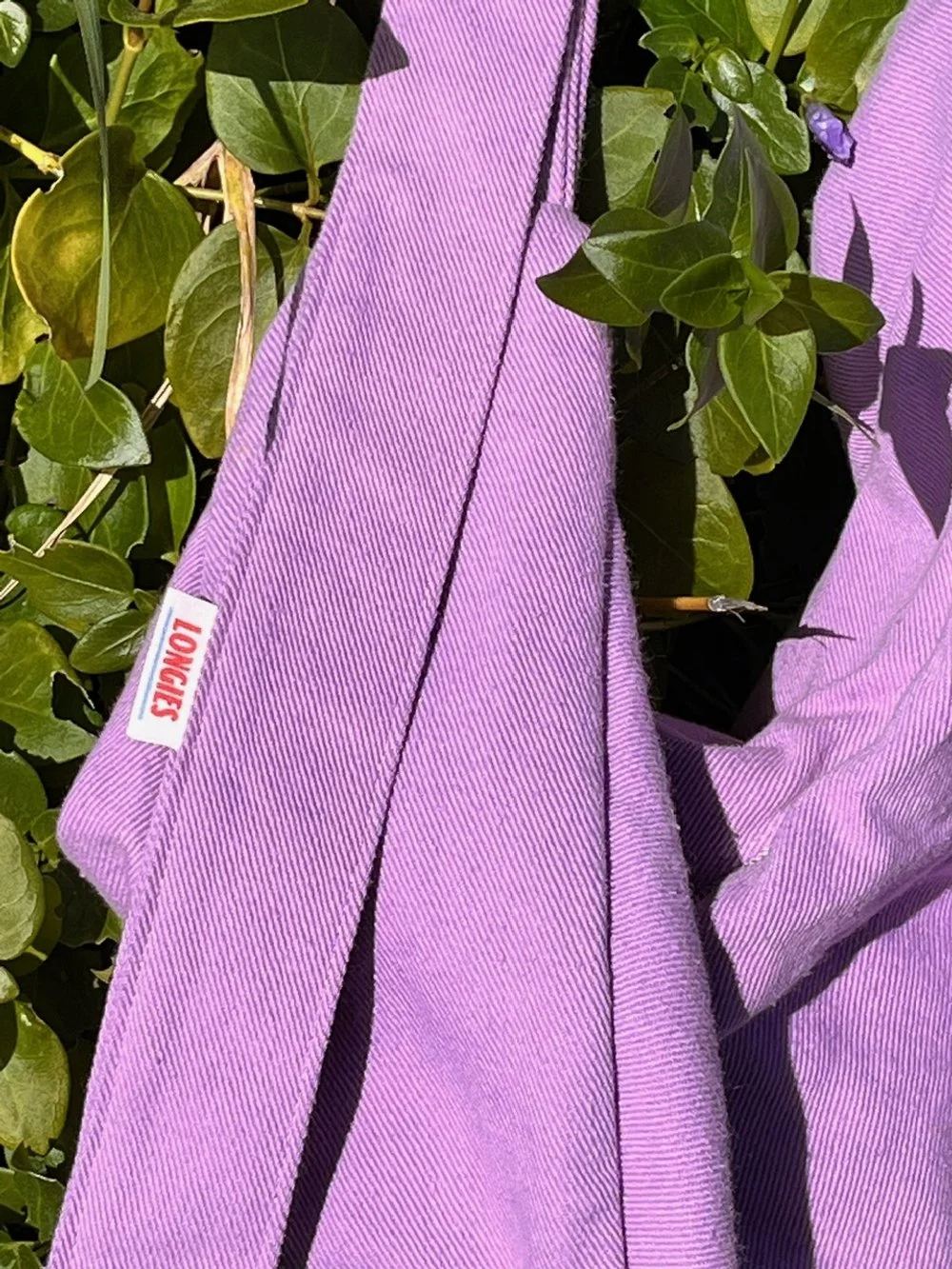 Close-up of a lupin lavender purple coloured denim Longines strap with a Longies tag hanging among green leaves.