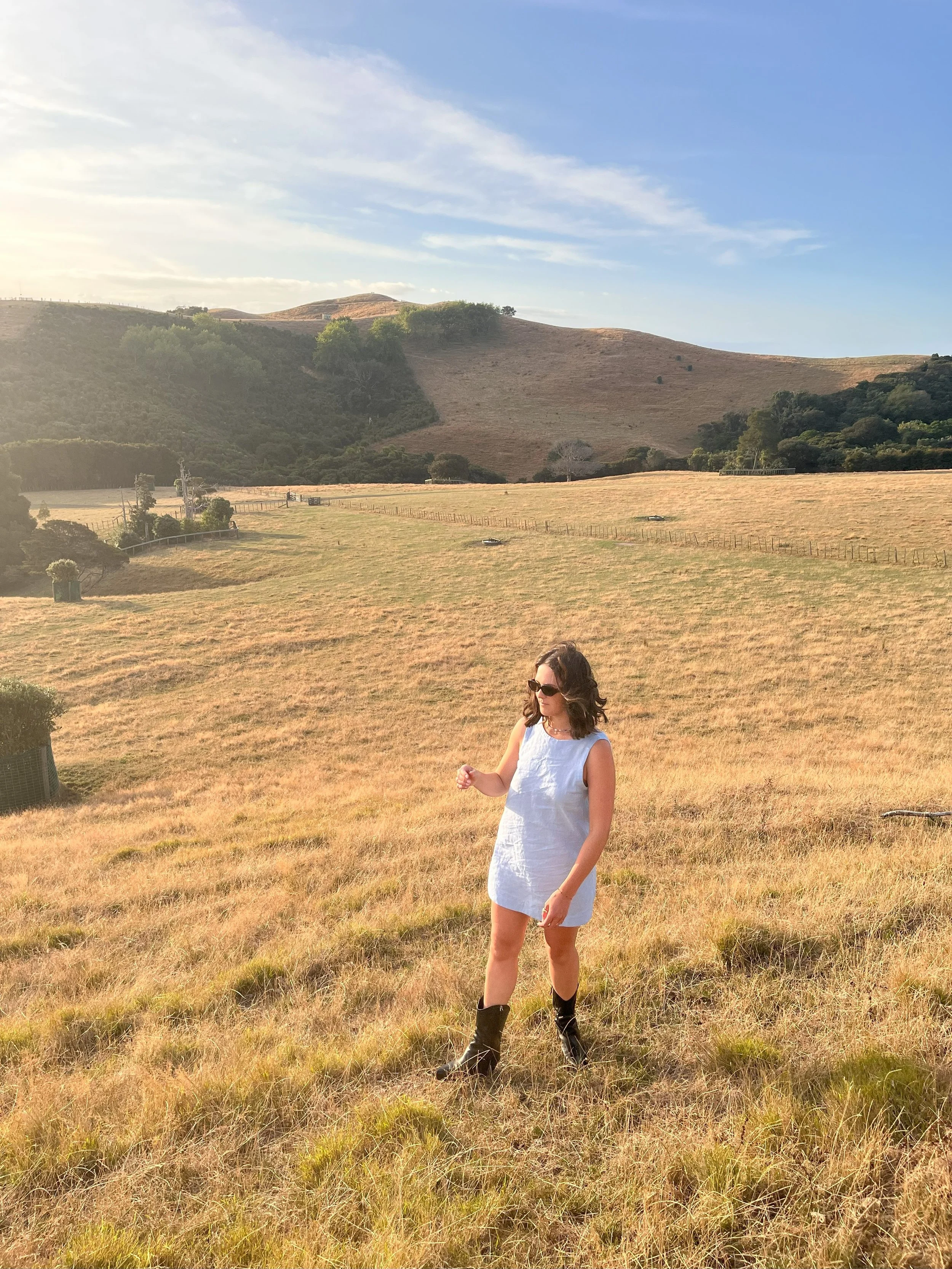 A woman in a blue a line sleeveless dress and sunglasses standing in a grassy field with rolling hills in the background during late afternoon sunlight.
