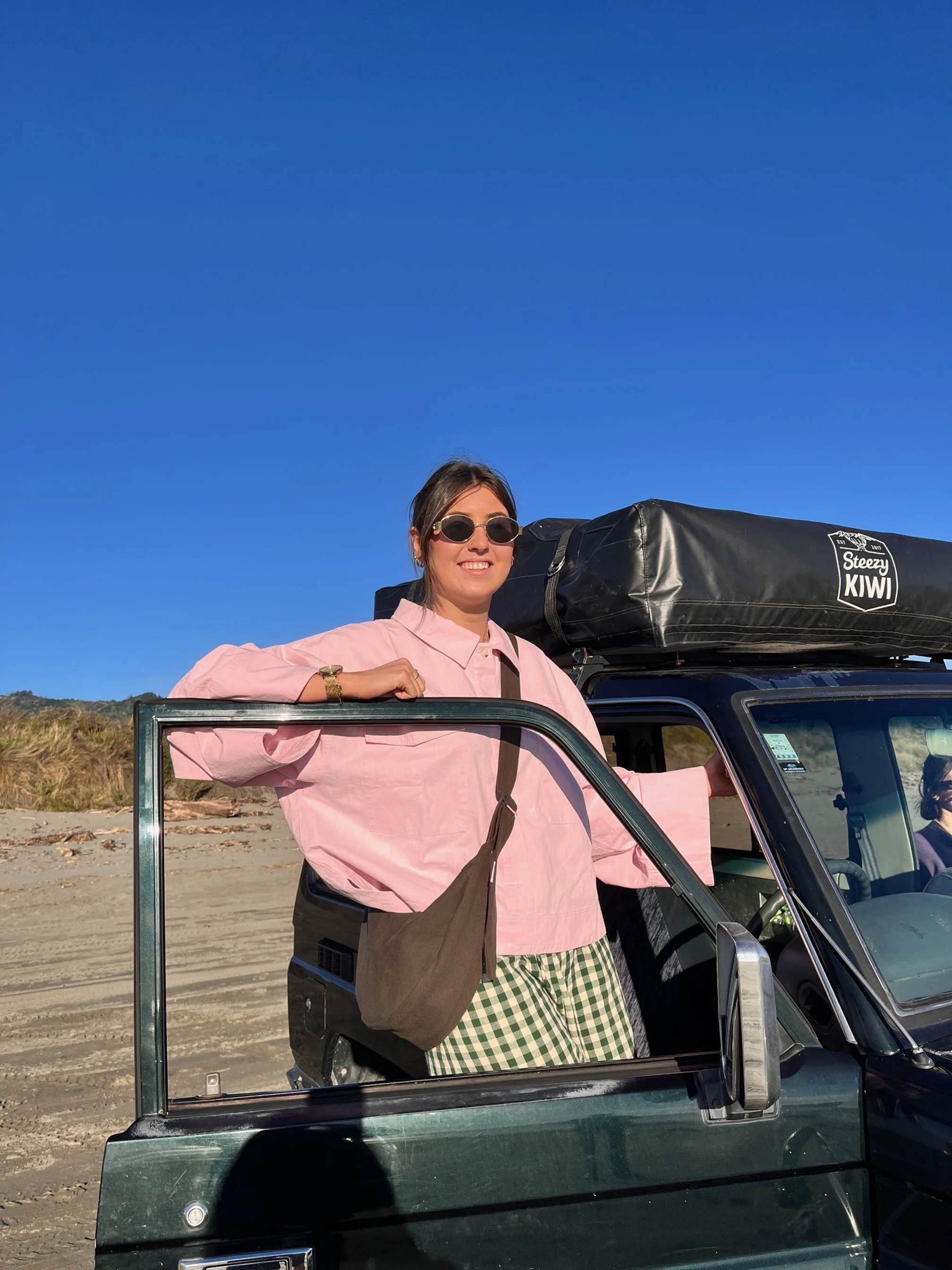 A woman wearing sunglasses, a pink jacket, and checkered pants, standing through the open door of a black off-road vehicle with a roof tent on a sandy beach landscape under a clear blue sky. Wearing her Tones collection cinderwood denim Longies bag.
