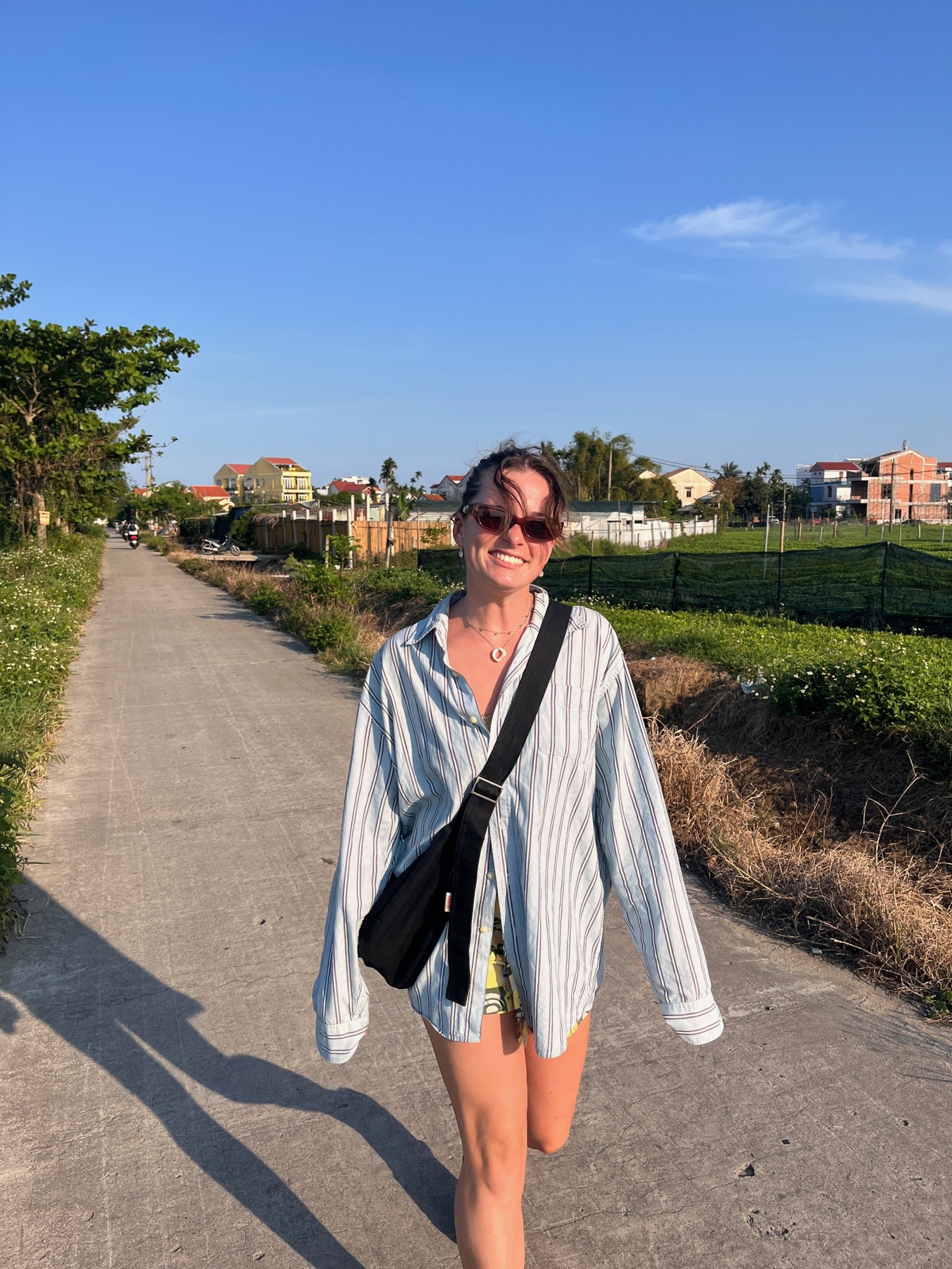 A woman wearing sunglasses, a striped shirt, and shorts walking outdoors on a sunny day, smiling, with a clear blue sky and houses in the background, in Vietnam wearing a Long Black, black handmade denim Longies bag.