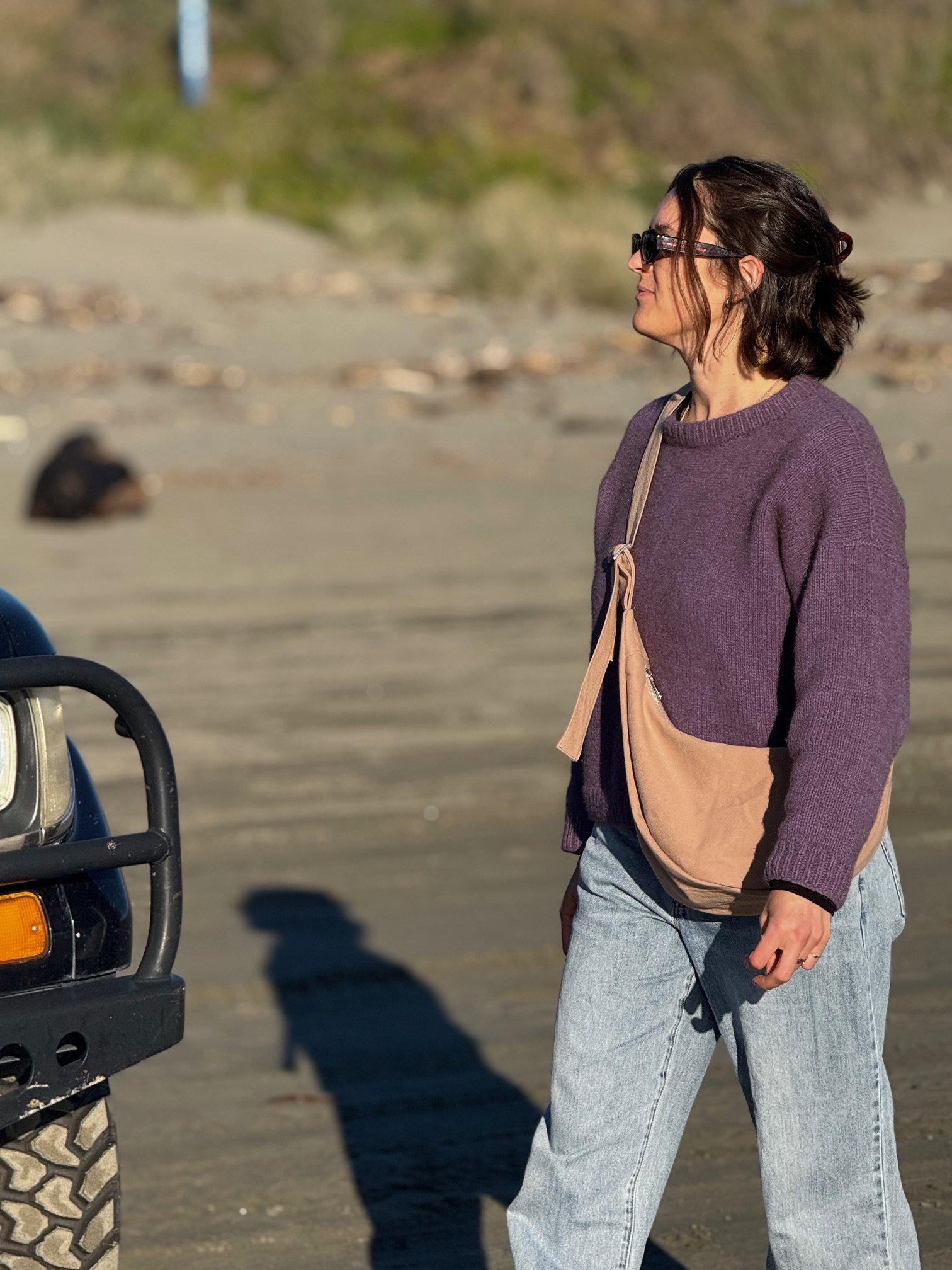 A woman with dark hair wearing sunglasses, a purple knit sweater, and light blue jeans standing on a beach, wearing the tone collection denim handmade Longies crossbody bag in dune, sand colour.