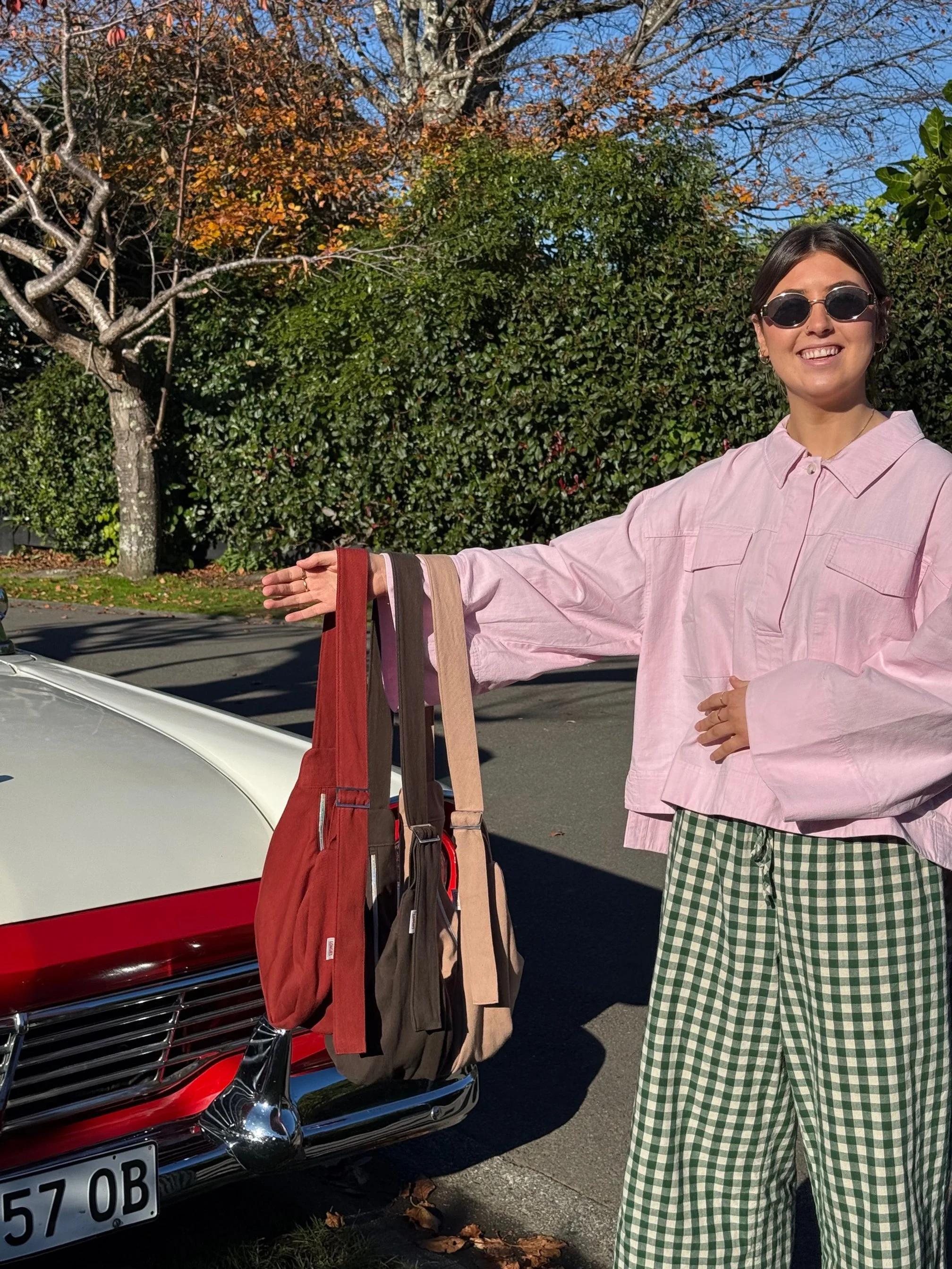 A woman standing next to a white and red car, holding the tones collection denim Longies bags. Ember red, dune sand, cinderwood brown She is wearing a pink oversized shirt.