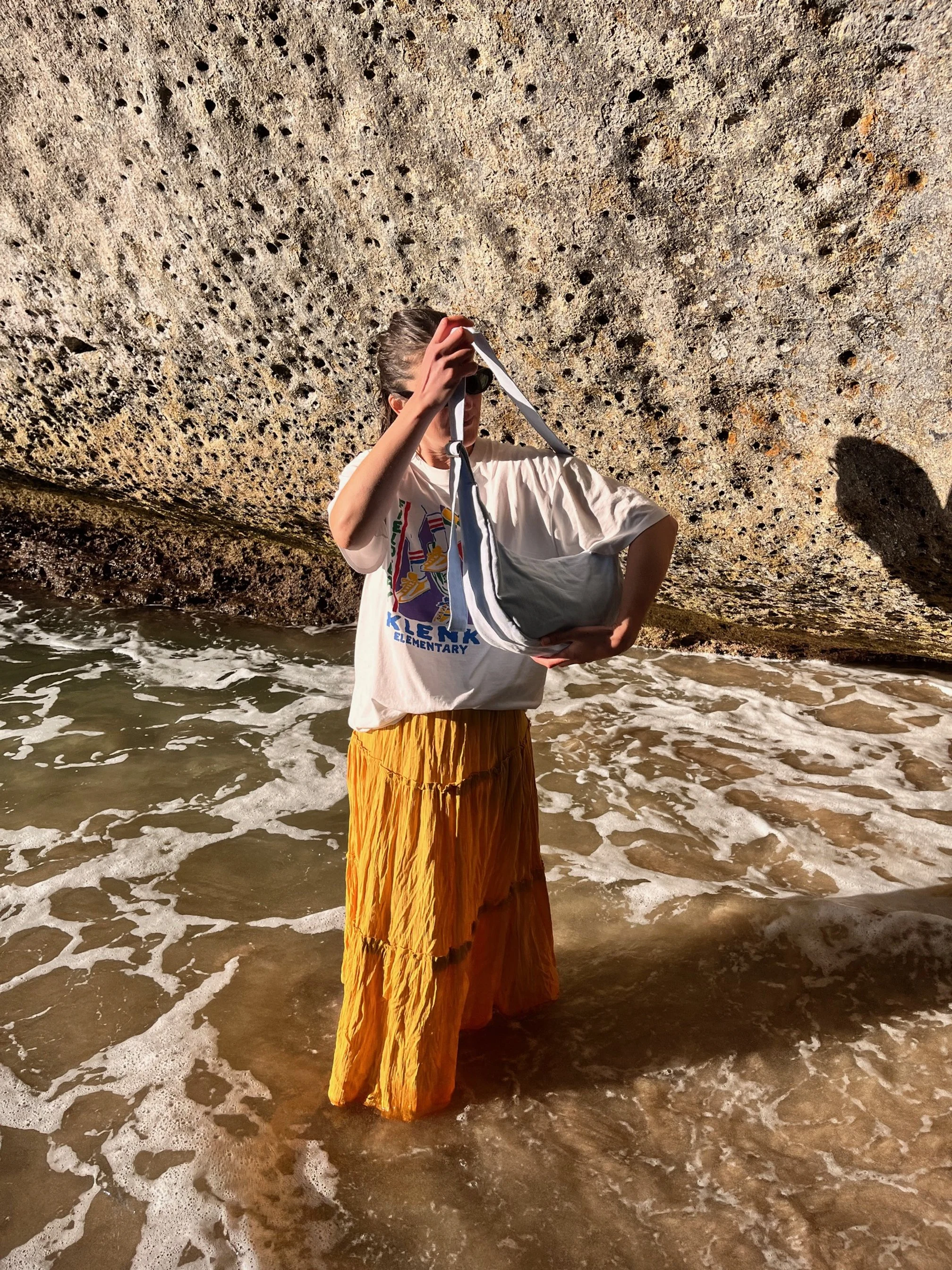 Person standing in shallow ocean water by a large rock wall, holding mist blue denim crossbody Longies bag, wearing a white T-shirt and yellow skirt.