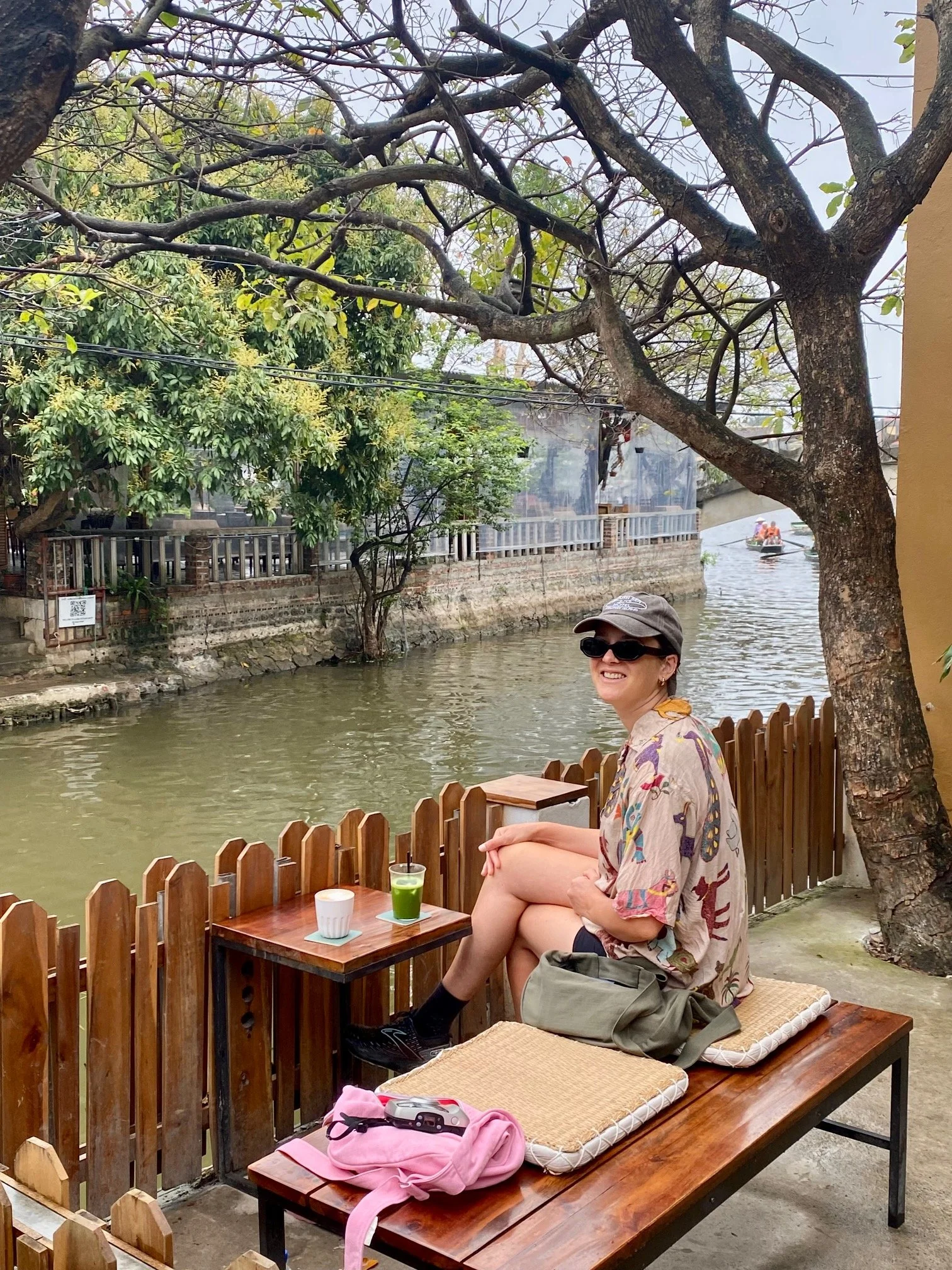 A woman wearing sunglasses, a hat, sitting on a bench by a waterway in Vietnam with a matcha in front of her and smiling, with trees and a small boat in the background. With denim Longies bags in peony pink and olive green, great for travelling.