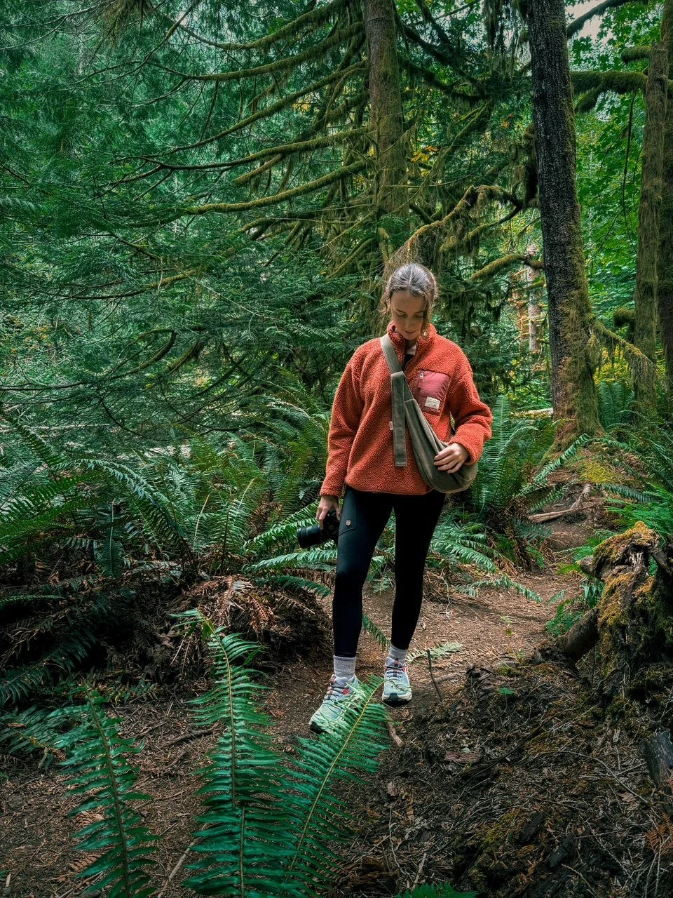 A woman in an orange fleece jacket and black leggings hiking on a wooded trail surrounded by green ferns and tall trees in Canada wearing a handmade denim Olive Longies corssbody bag.