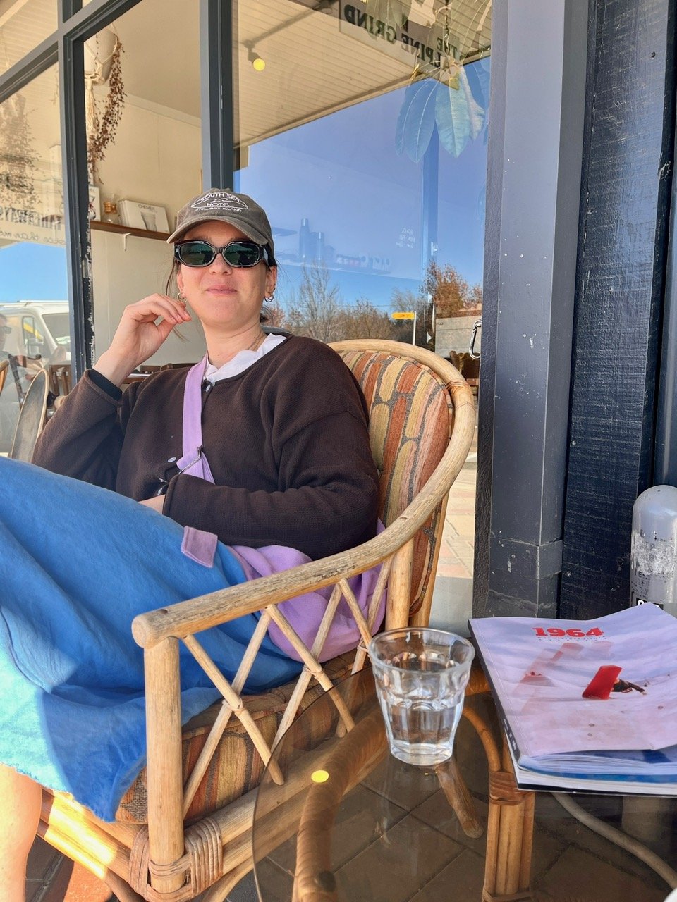 A woman wearing sunglasses, a gray baseball cap, a black jacket, and blue pants sits on a wicker chair outside a cafe, with a glass of water and a magazine on a glass-top table nearby, wearing her orchard collection Lupin crossbody Longies bag.