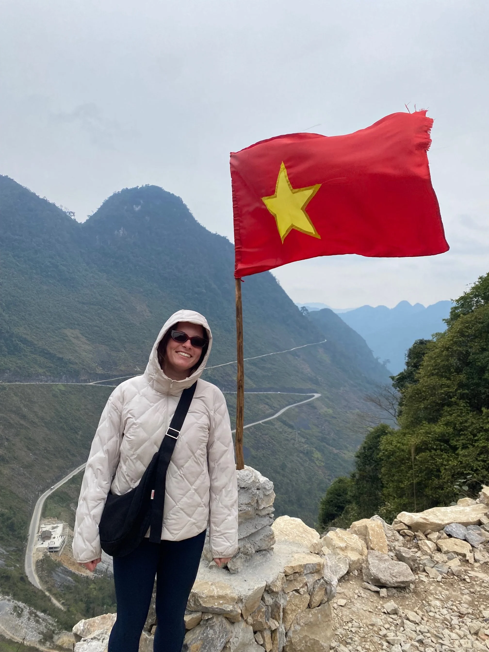 A woman wearing her great traveling crossbody Longies bag in black denim, smiling, stands next to a flag of Vietnam on a mountain trail. The background features green, mountainous terrain with winding roads under an overcast sky.