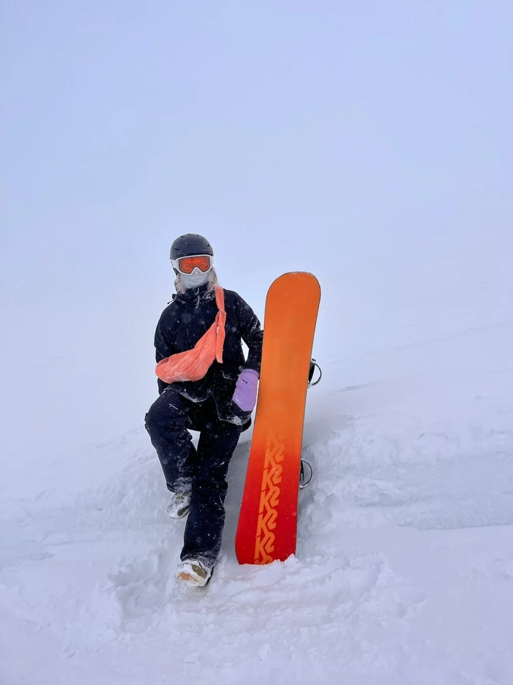 Person wearing ski gear, including helmet, goggles, and face mask, sitting or kneeling next to orange snowboard on snow in a foggy or snowy environment. wearing her peach orange coloured handmade denim Longies bag.
