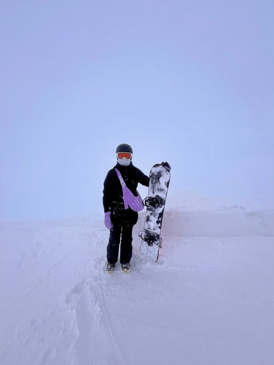 Person in black snowboarding gear, wearing a helmet and goggles, standing in the snow with a snowboard upright next to them on a foggy, snowy landscape, wearing orchard collection crossbody denim Longies bag in the colour lupin purple.