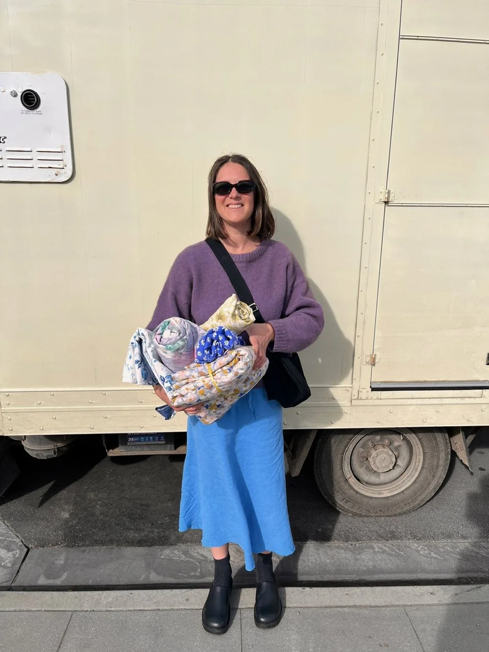 Woman smiling with dark sunglasses, purple sweater, blue skirt, black shoes, holding deadstock repurposed fabric to make handmade denim crossbody Longies bags, while travelling in a truck around NZ.
