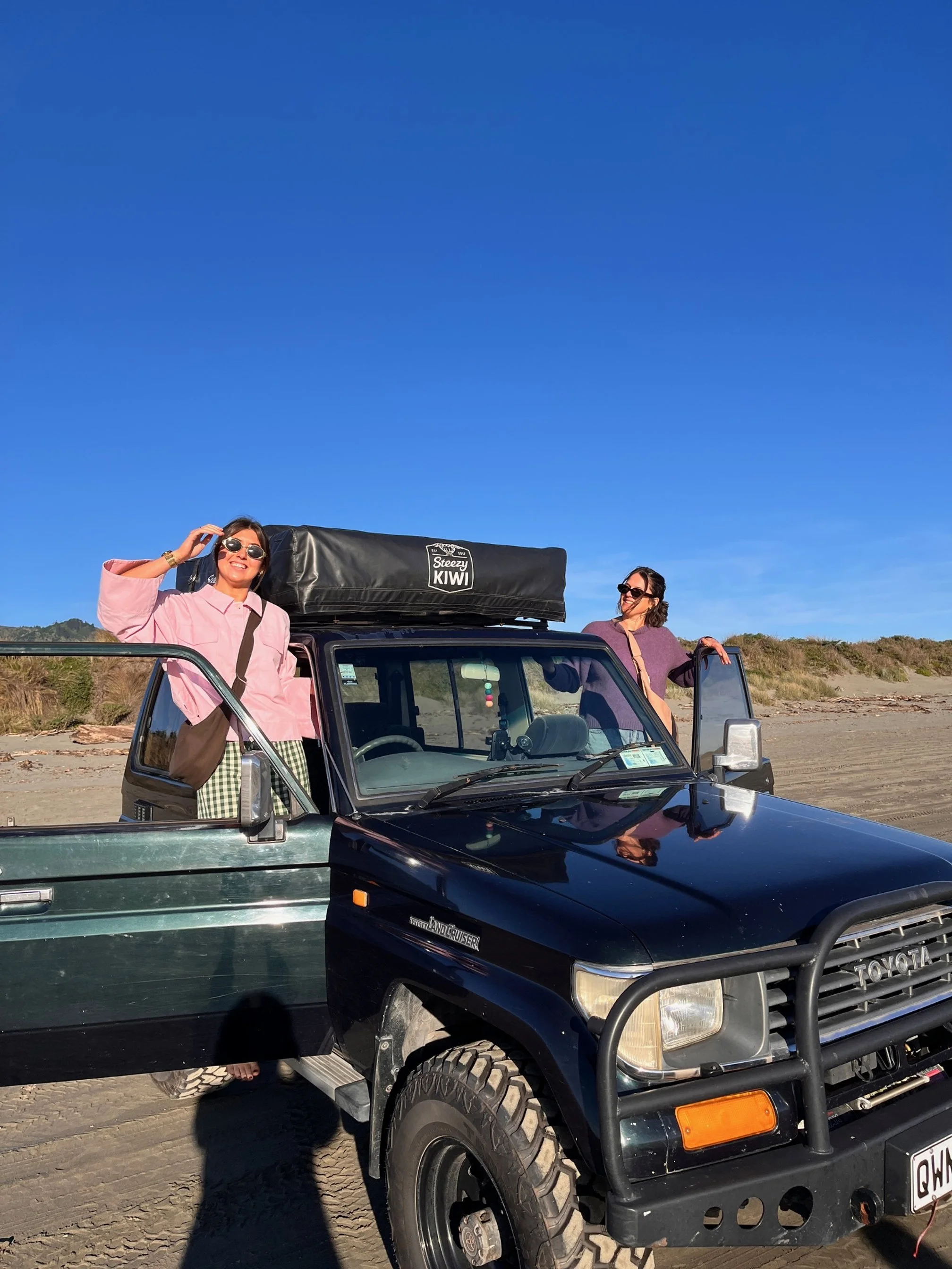 Two women standing outside a black Toyota Land Cruiser on a sandy beach, with a clear blue sky in the background. Wearing their tones collection handmade denim crossbody Longies bags in cinderwood brown and dune sand colours.