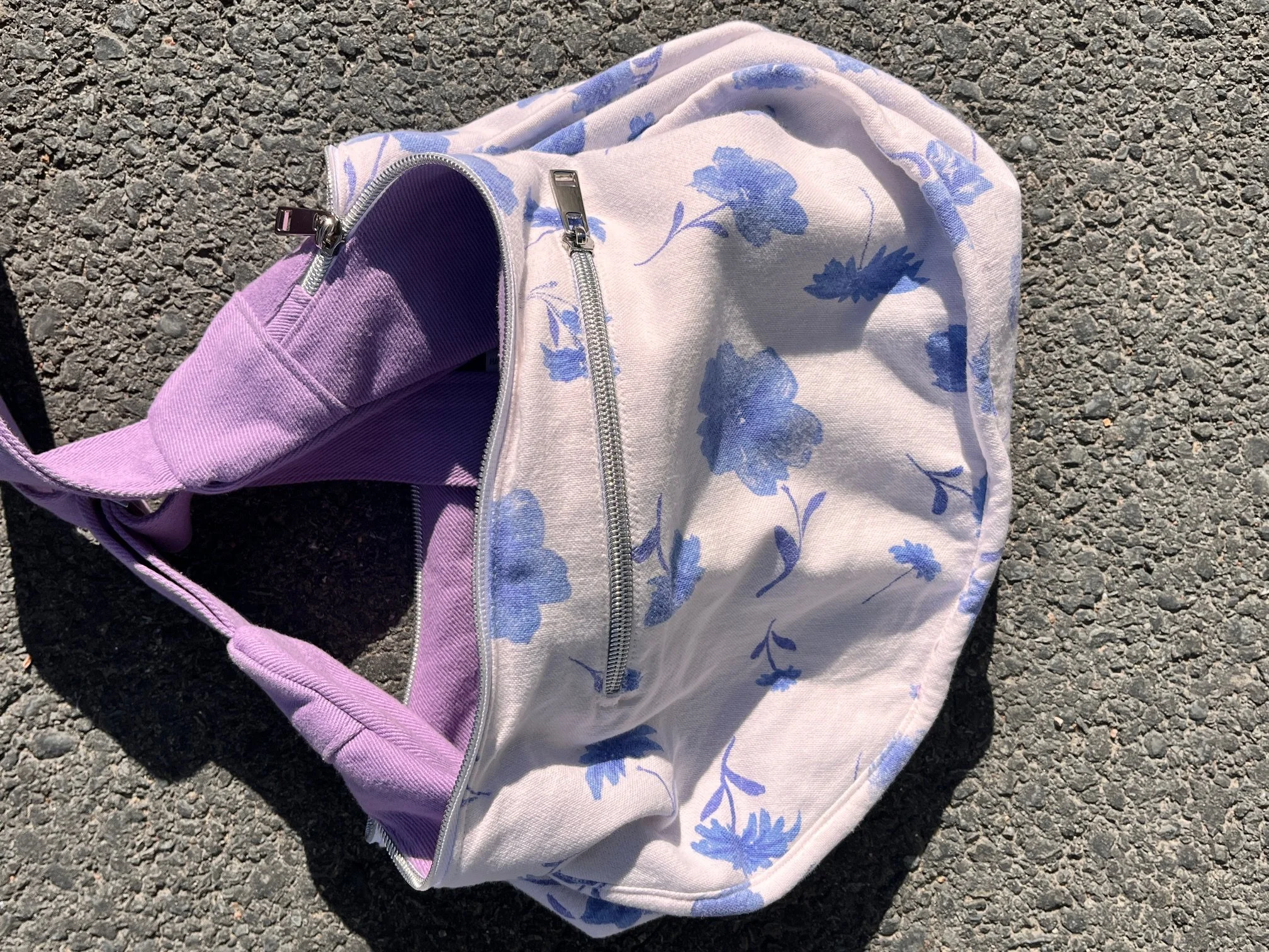 A Lupin purple coloured bag from the new Longies orchard collection, with white and blue floral pattern upcycled lining and a zipper pocket, lying on an asphalt surface.