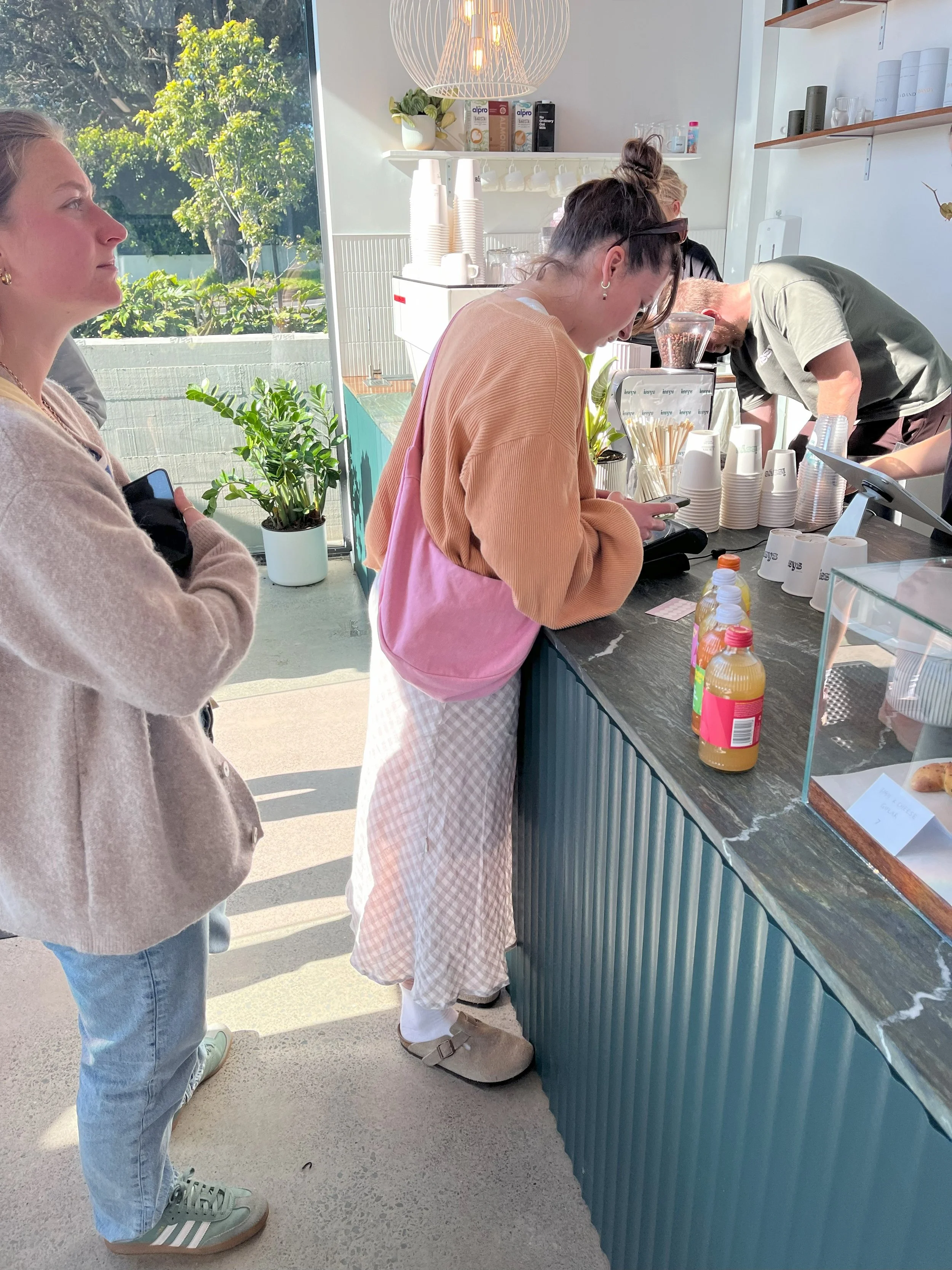 A woman with a beige sweater and blue jeans standing in line at a cafe, while another woman in an orange and pink sweatshirt and checkered pants places her order at the counter. With a handmade denim peony pink Longies bag over her shoulder.
