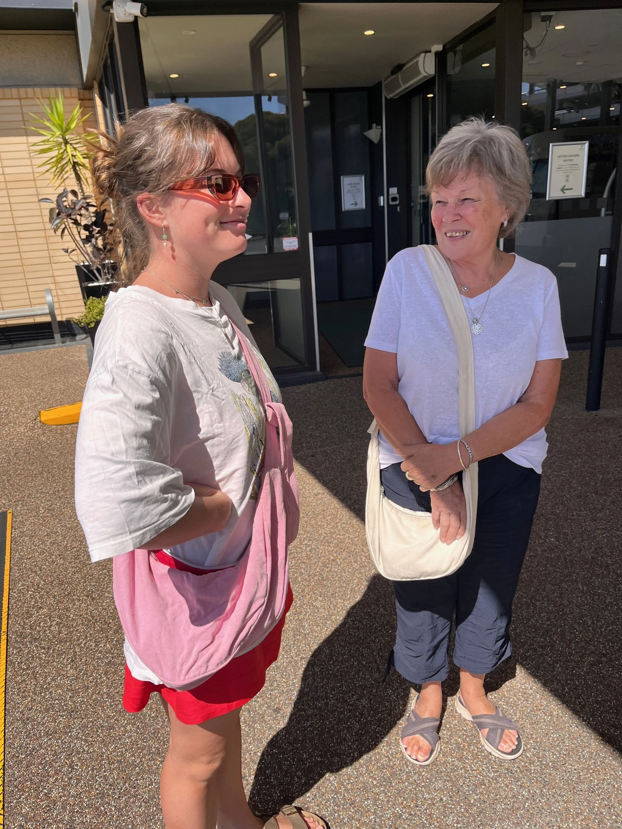 Two women are standing outdoors near the entrance of a building, engaged in conversation. The woman on the left wears sunglasses, a white T-shirt, red shorts, and carries a pink bag. The woman on the right has short gray hair, wears a white T-shirt, 