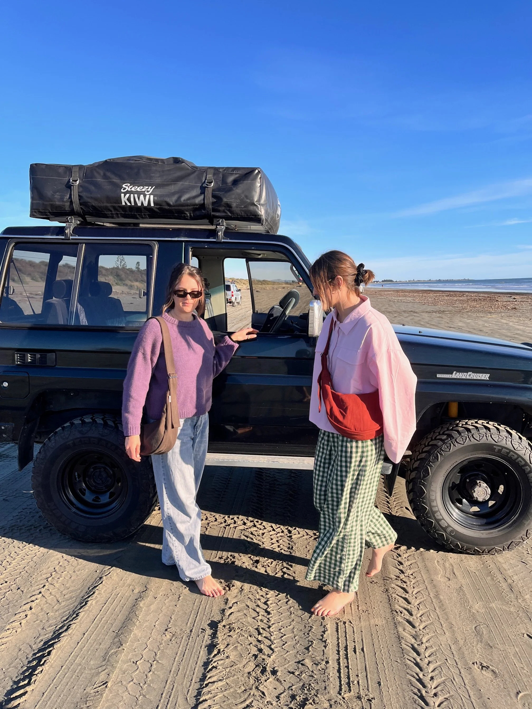 Two women standing barefoot on the sand near a black off-road vehicle with a roof cargo bag on the beach, with the ocean and a clear blue sky in the background. Wearing the tones collection handmade denim crossbody Longies bags.