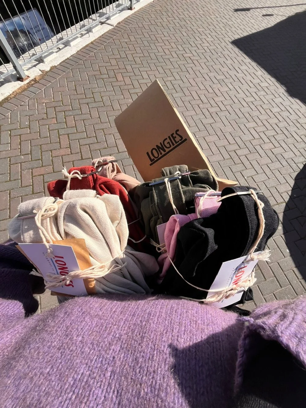 A person holding parcels of Longies bags in brown cardboard packaging ready to send to customers black, pink, beige, red, and green colors, with a cardboard box labeled 'LONGIES' on a brick sidewalk near a railing.