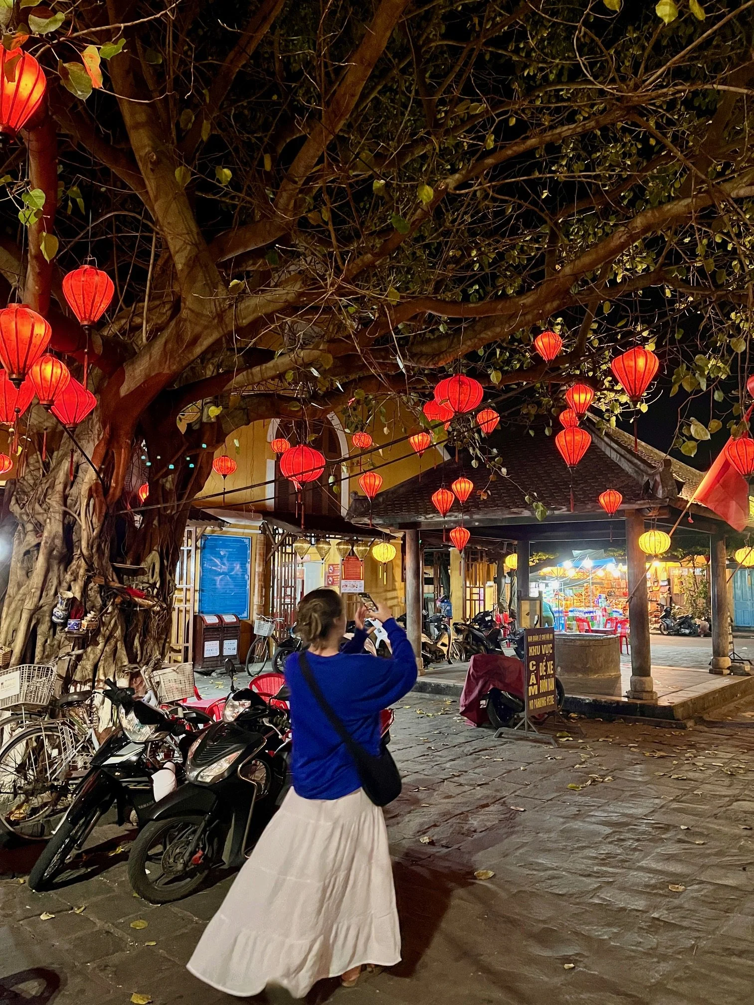 A woman in a blue top and white skirt takes a photo outdoor area at night decorated with red lanterns hanging from trees. Wearing her long black, black coloured denim handmade Longies bag made in NZ.