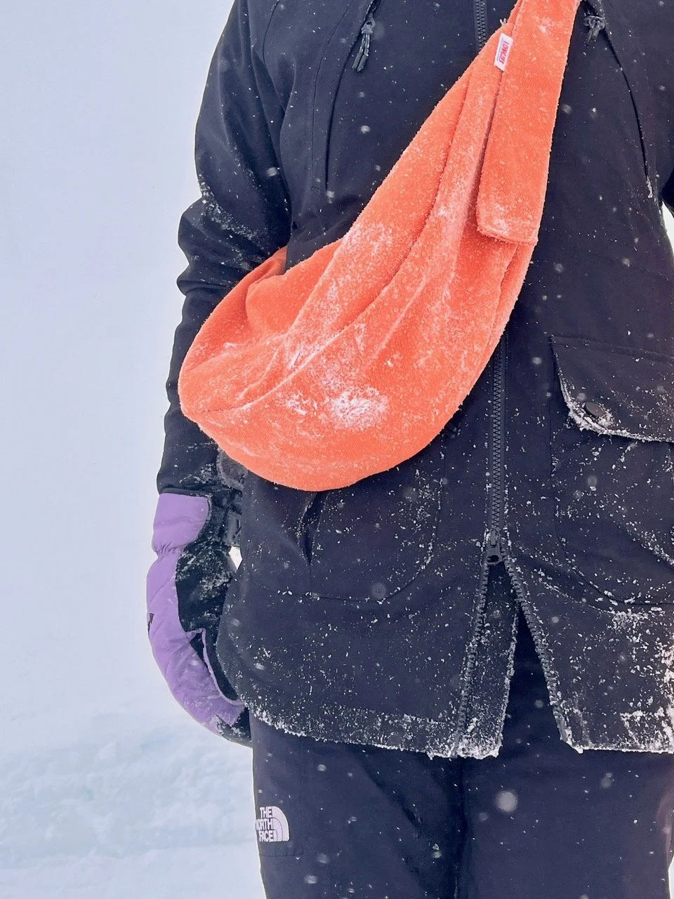 Person wearing a black jacket and black pants, holding a peach orange denim handmade Longies crossbody bag over their shoulder, standing in snow with snowflakes around.