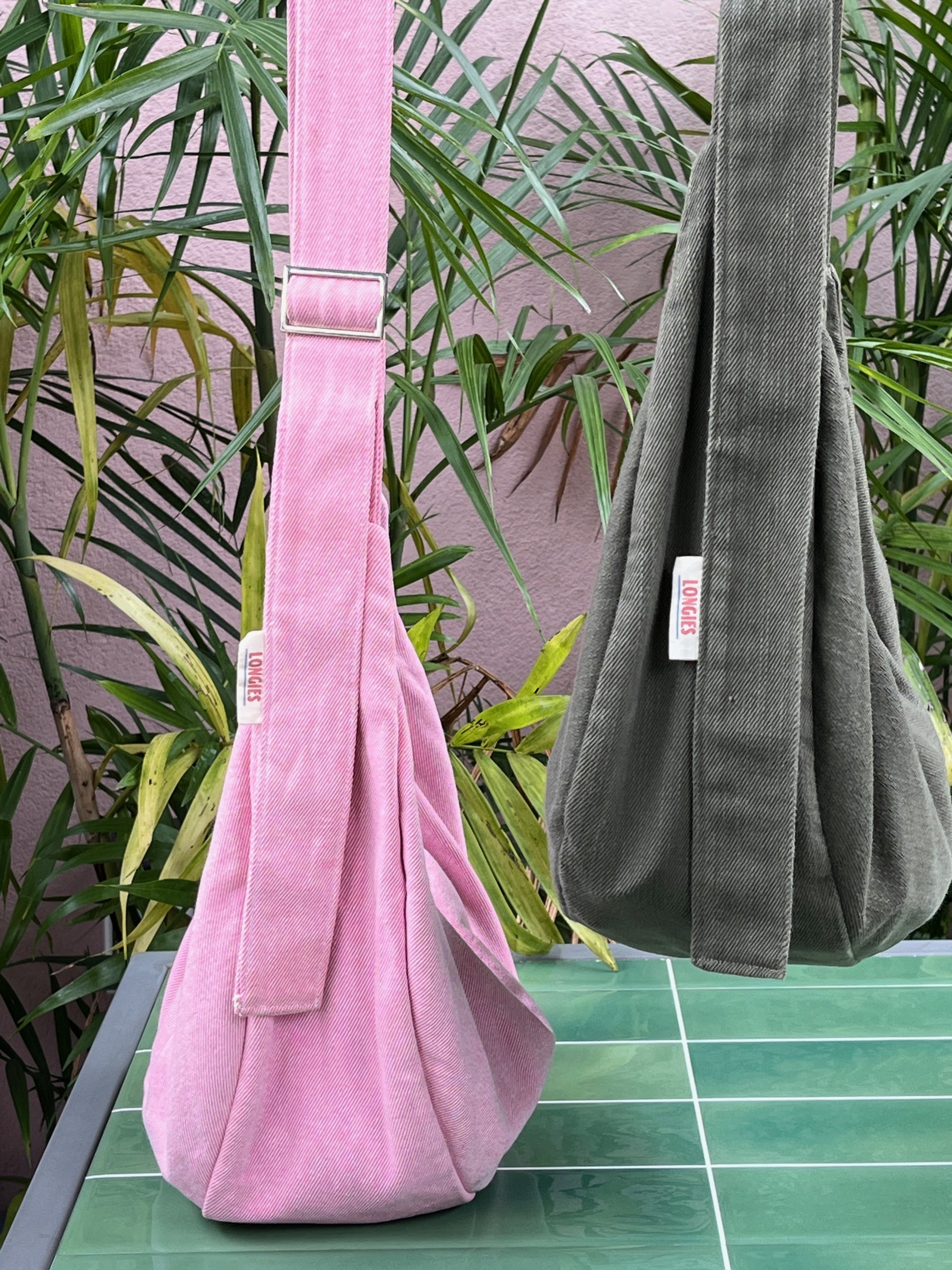 Two handbags hanging, one is peony pink and one is olive green, with tags that say 'LONGIES'. They are handmade denim crossbody bags made in NZ. The background is a pink wall, and the surface beneath the bags is green tiled.