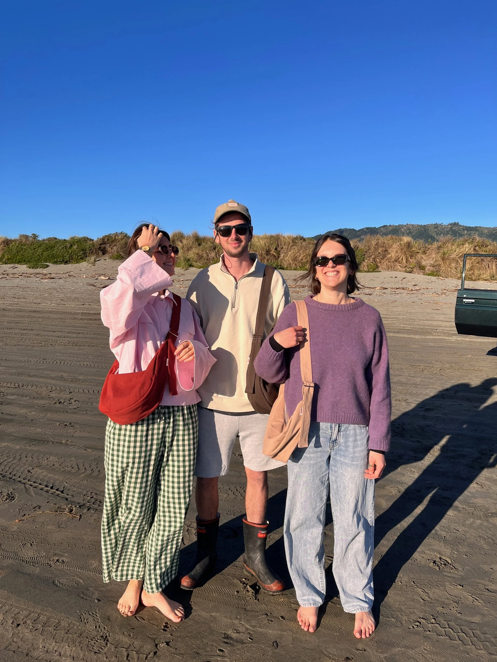 Three people standing on a sandy beach, smiling and wearing sunglasses, with dunes and a clear blue sky in the background.