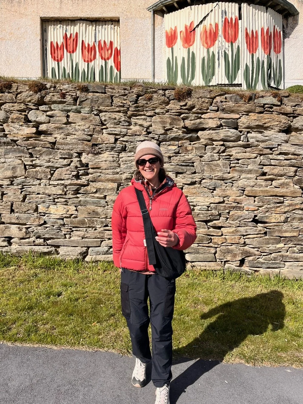 A woman in a red puffer jacket, black pants, beige beanie, a Longe black crossbody Longies bag and sunglasses is smiling and standing outdoors on a paved path with a stone wall behind her. Above her are painted tulip flowers on two small windows.