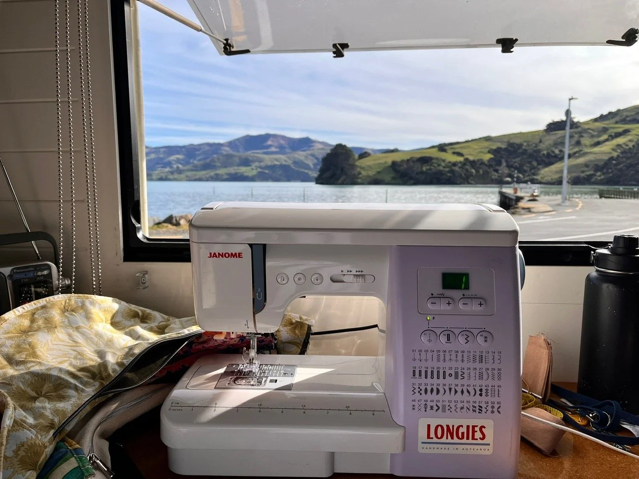 A sewing machine on a table near a window showing a landscape with water, mountains, and greenery outside. Sewing while travelling on the road in New Zealand.