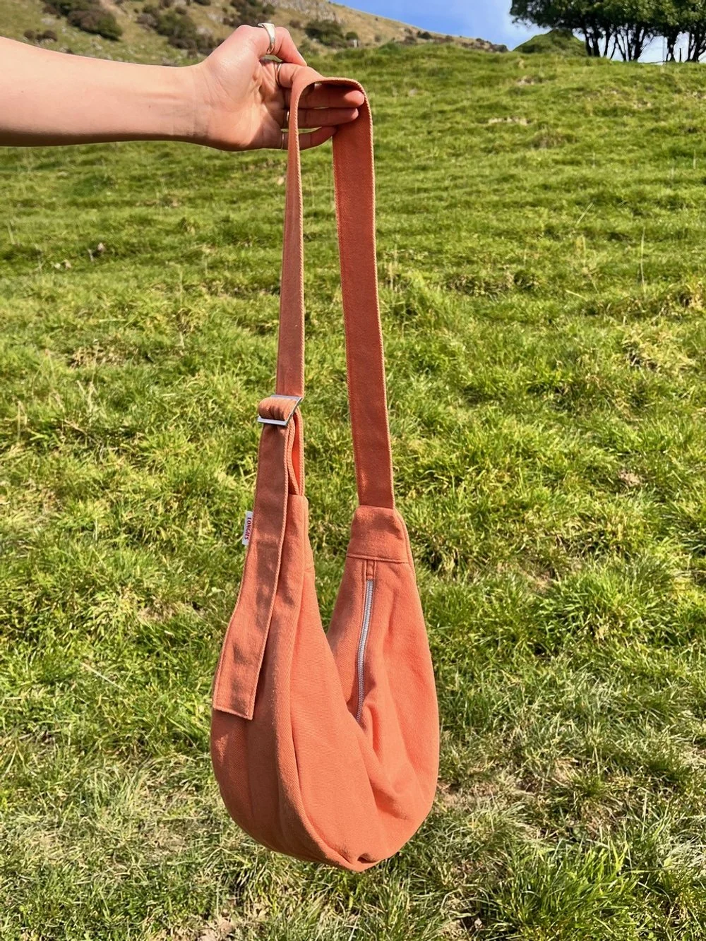 A person's hand holding an orchard collection peach orange handmade denim Longies crossbody bag with a long strap in an outdoor grassy area under a blue sky.