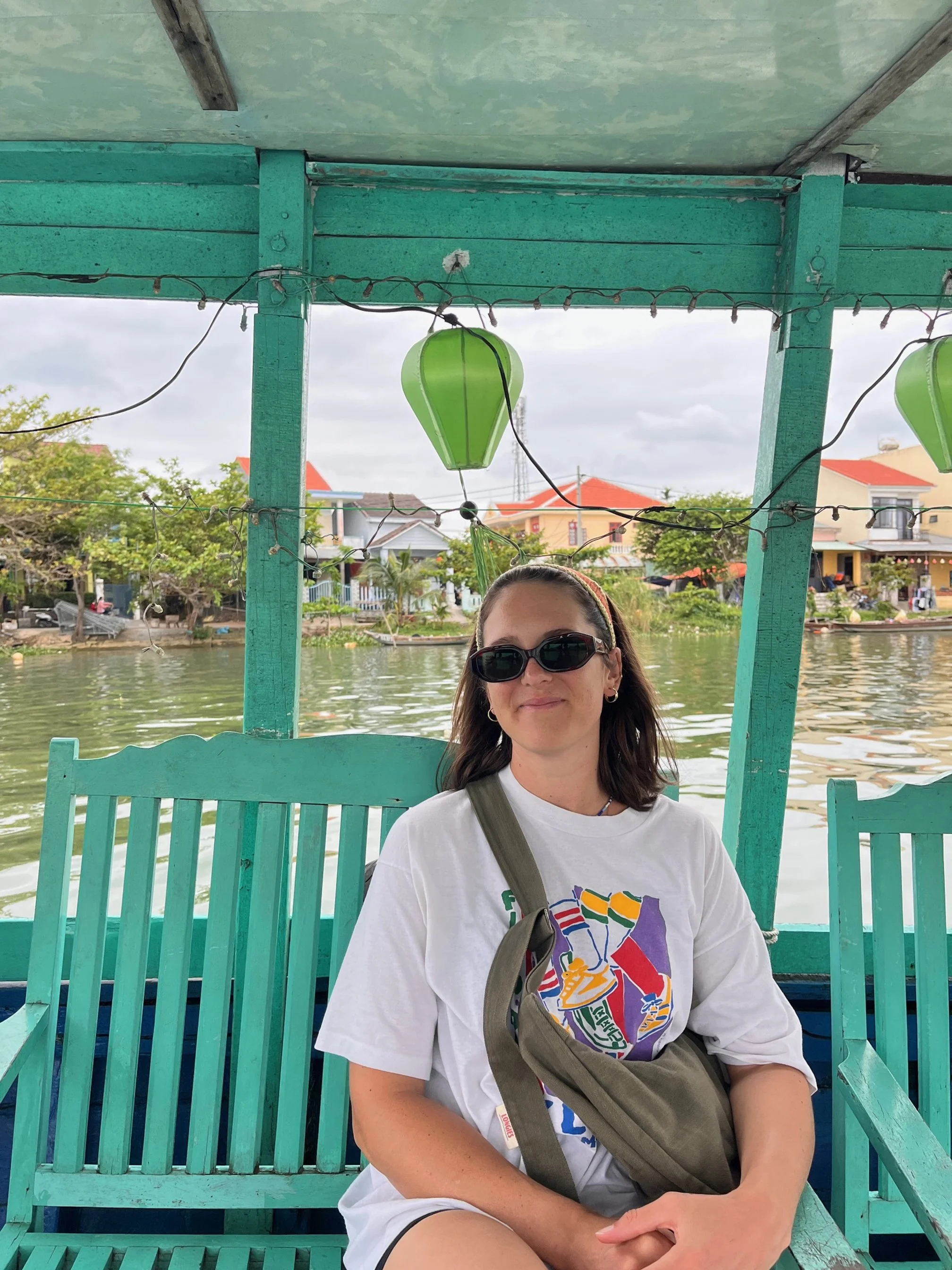 A woman sitting on a teal wooden bench in a boat on a river, wearing sunglasses, a white graphic t-shirt, and a olive denim crossbody Longies bag, with houses and trees in the background and green lanterns hanging above.