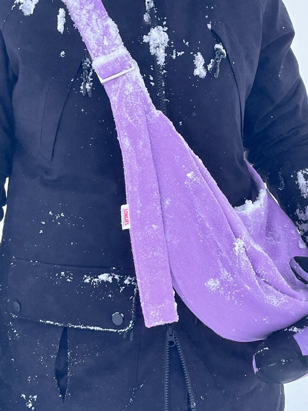 Close-up of a person wearing a black winter jacket and purple gloves, covered in snow, wearing orchard collection crossbody denim Longies bag in the colour lupin purple.