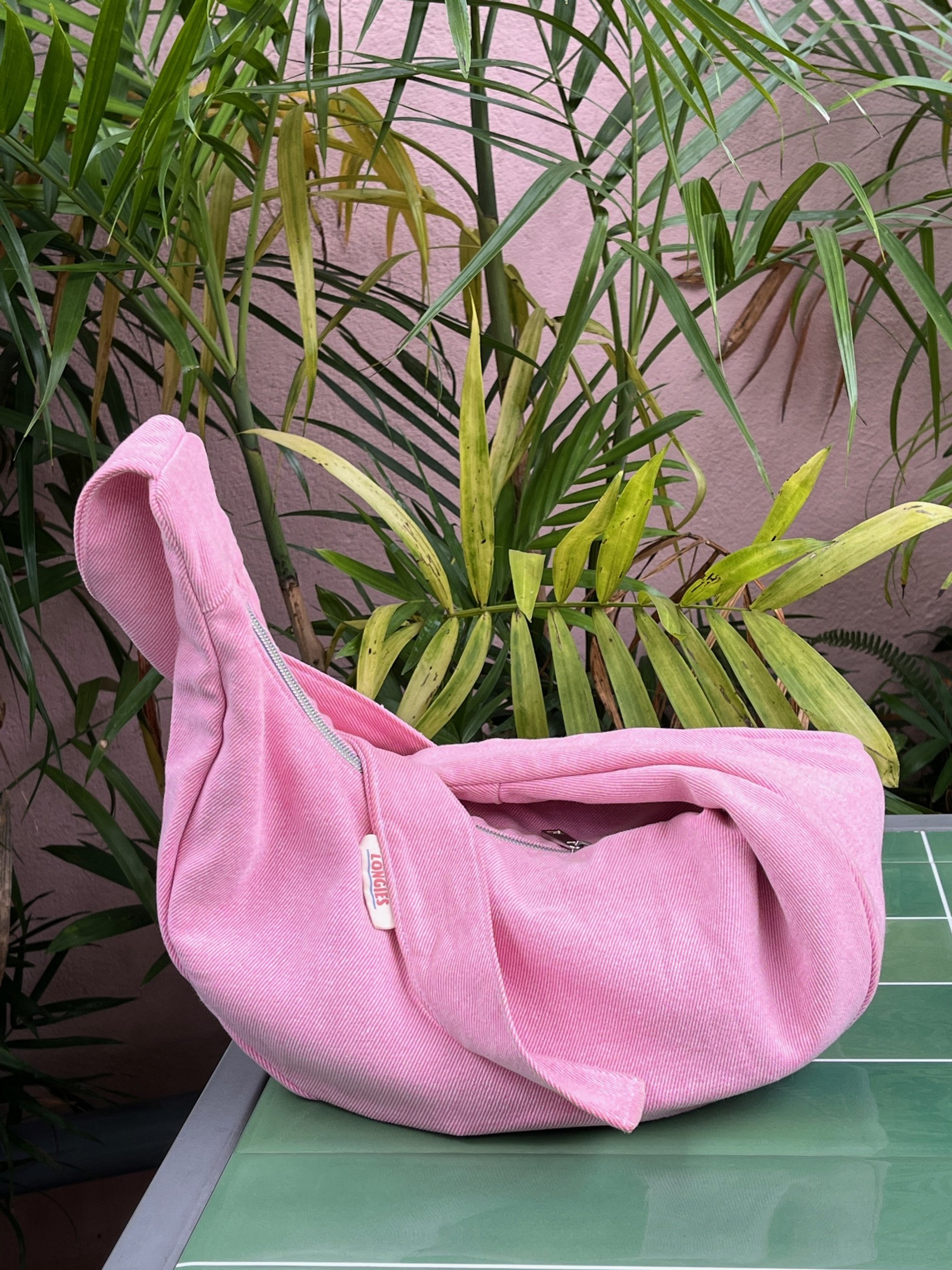 Peony pink handmade denim Longies bag with Zippers on a Green Tile Table, surrounded by green tropical plants in a pink-walled outdoor setting in Vietnam.