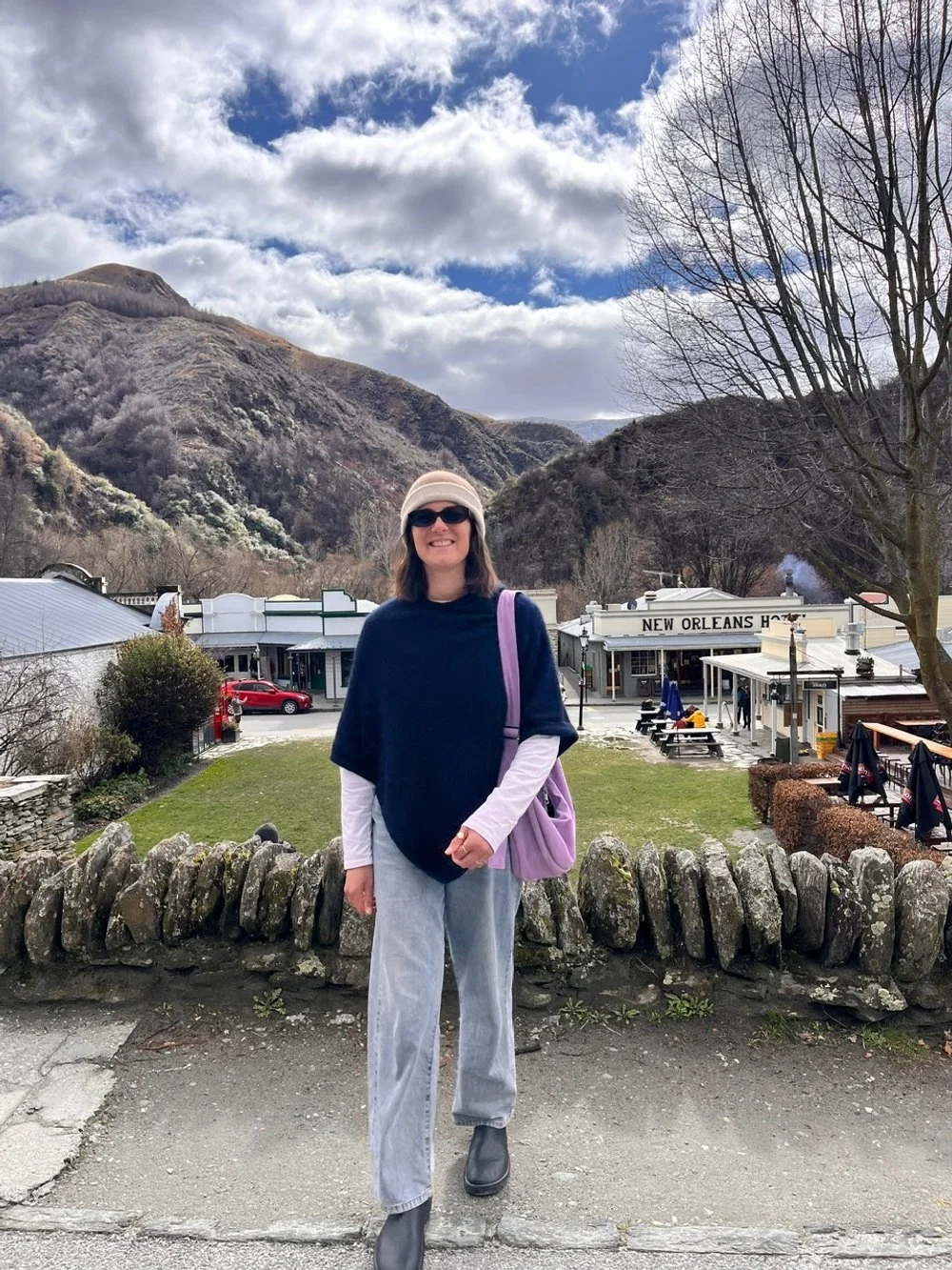A woman standing outdoors in front of a small town with mountains in the background, wearing sunglasses, a beige beanie, a dark blue poncho layered over a white long-sleeve shirt, gray pants, black shoes, and carrying a handmade Longies purple bag.
