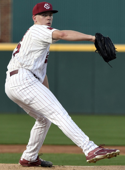A young baseball player in a white pinstripe uniform and red cap pitching on the field.
