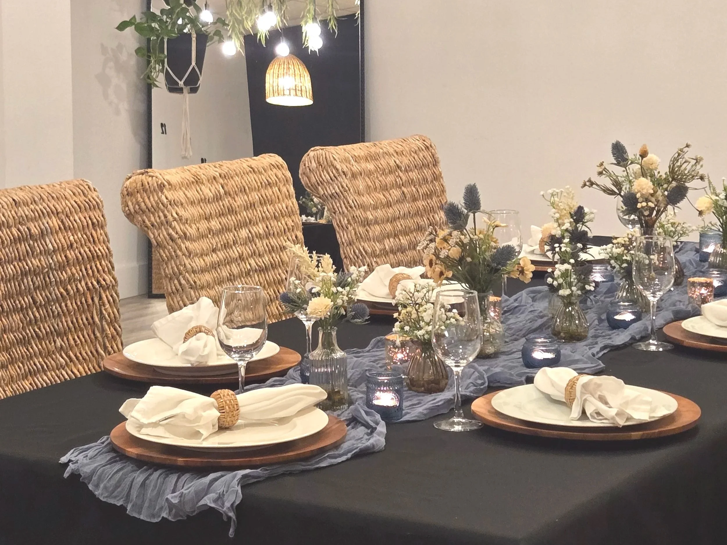 Blue table scarf with wooden chargers, white plates, goblets, flowers, wicker chairs, black tablecloth.