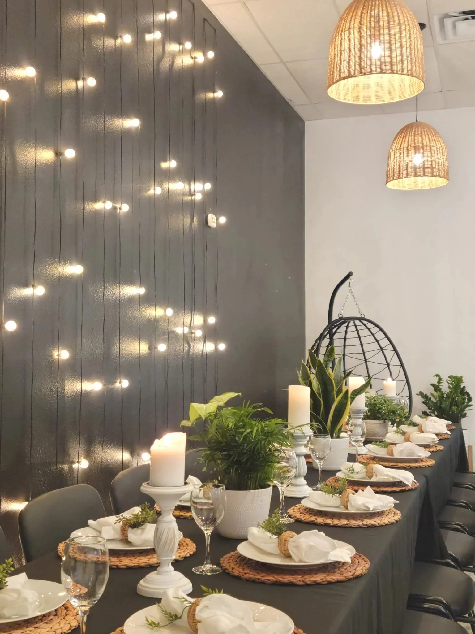 A dinner table set for a celebration with candles, potted plants, and elegant place settings, with decorative hanging wicker lamps and a string of fairy lights on a dark accent wall. Bridal Shower setup in our venue at Oasis Events Collective in Sara