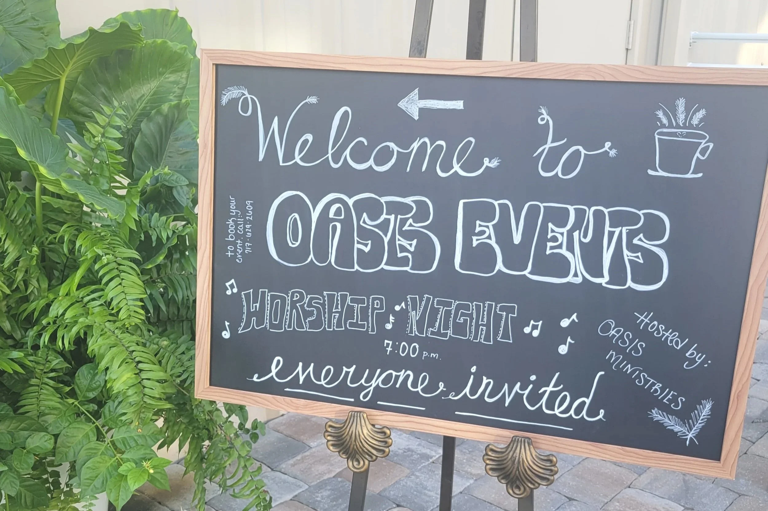 Chalkboard sign welcoming guests to a church event, with decorative drawings of a arrow, a teacup with plants, music notes, and text about worship night hosted by Oasis Ministries at 7:00 PM.