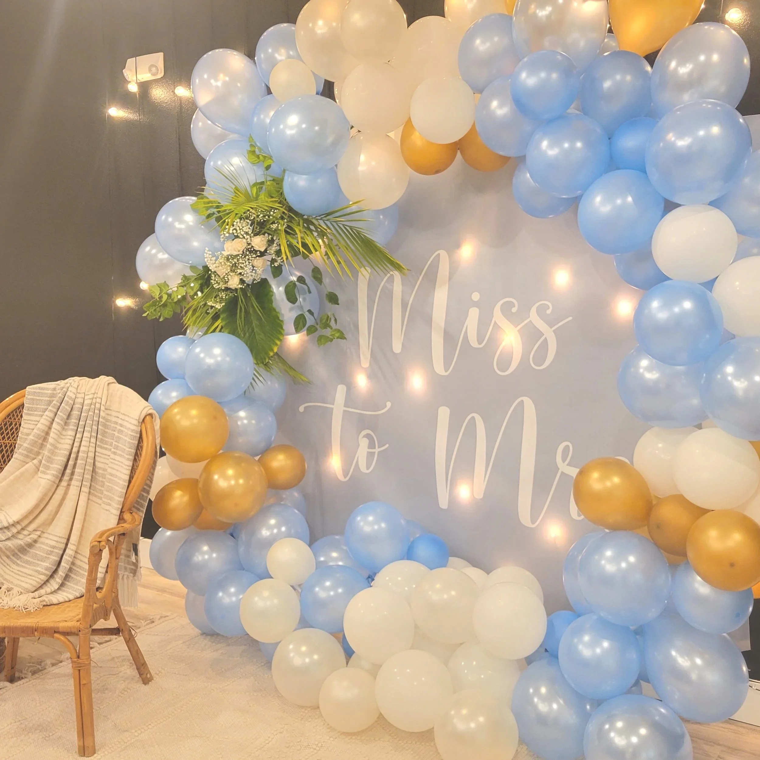 Decorative backdrop with blue, white, and gold balloons surrounding a white board that reads 'Miss to Mrs'. There are string lights and a floral arrangement on the left side.