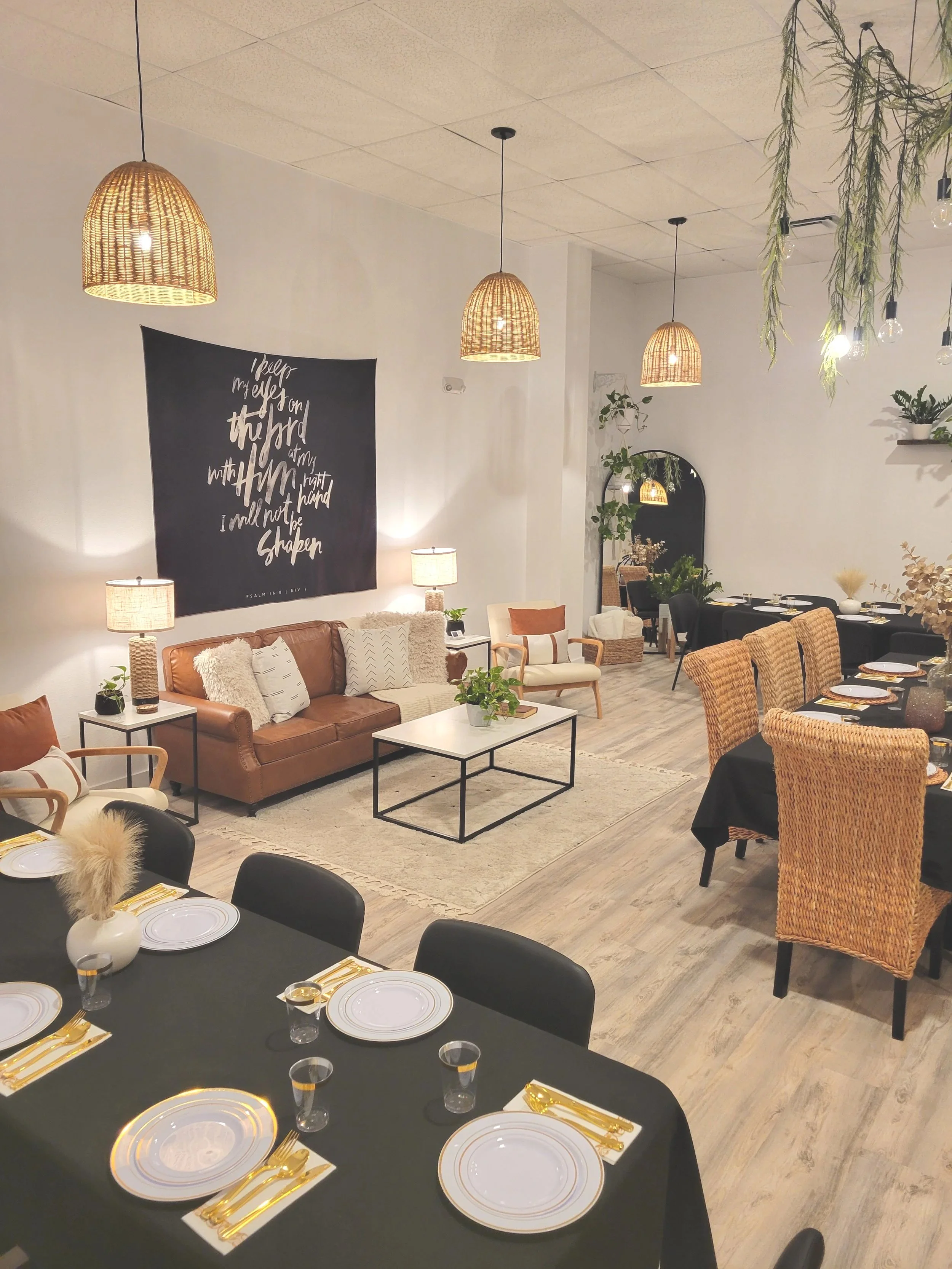 A cozy living room and dining area decorated with hanging pendant lights, a black table set with white plates and gold utensils, a leather sofa with pillows, a coffee table, and a dining table with rattan chairs, decorated with greenery and warm ligh