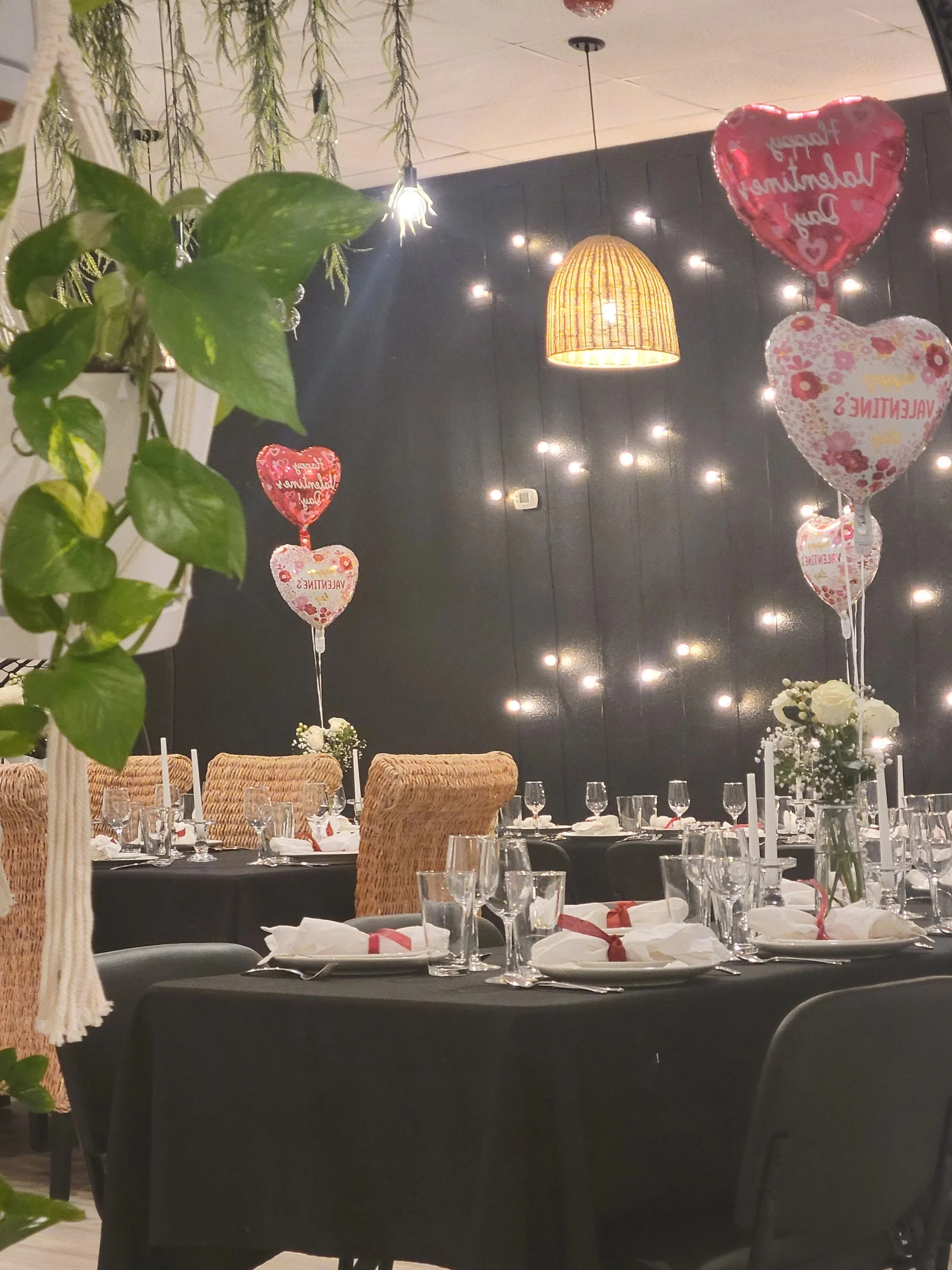 Valentine's Day themed dinner setup with tables, chairs, balloons, floral centerpieces, candles, glasses, and cutlery in a decorated room with string lights and hanging decor.