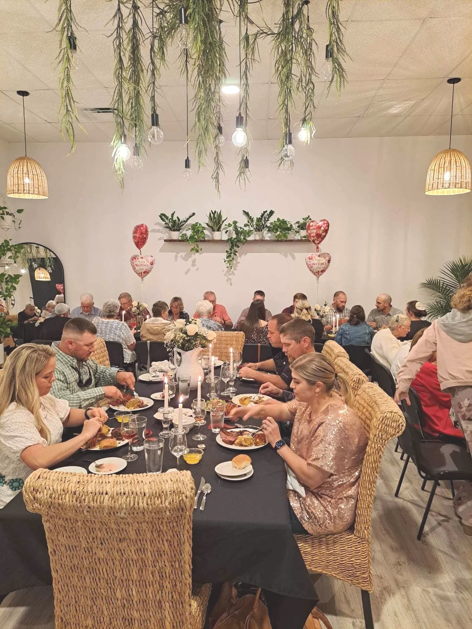 People dining at a banquet table during a celebration, decorated with balloons, candles, and floral arrangements, with greenery and hanging lights in a cozy, modern restaurant setting.