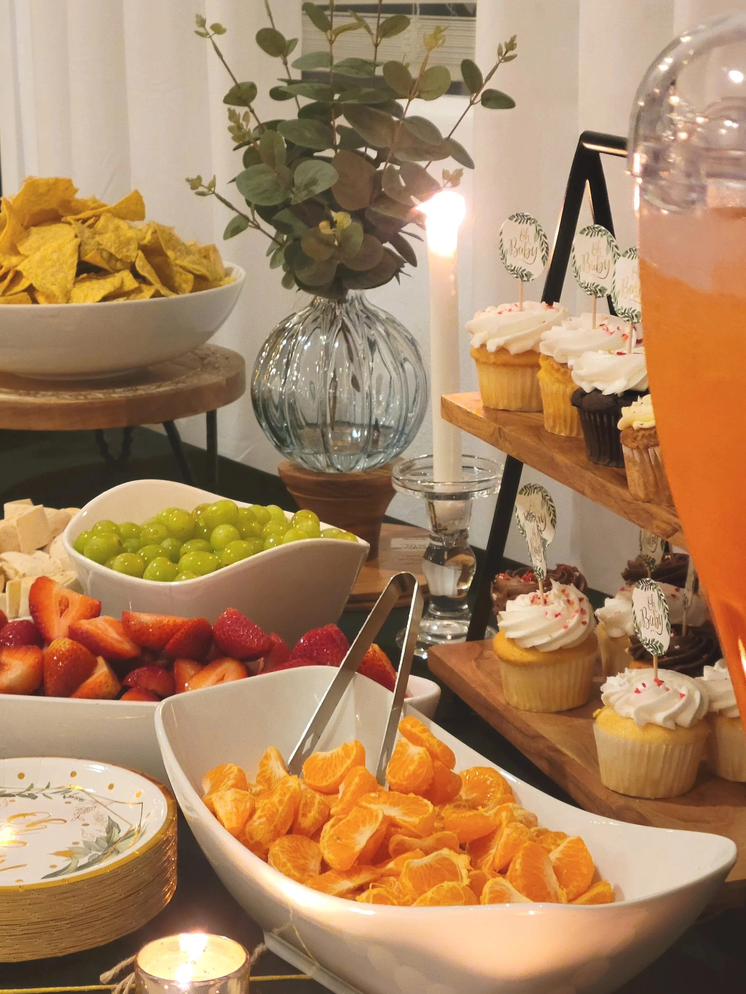 Assorted fresh fruit and cupcakes on a dessert table at a celebration, with a candle and flower arrangement in the background.