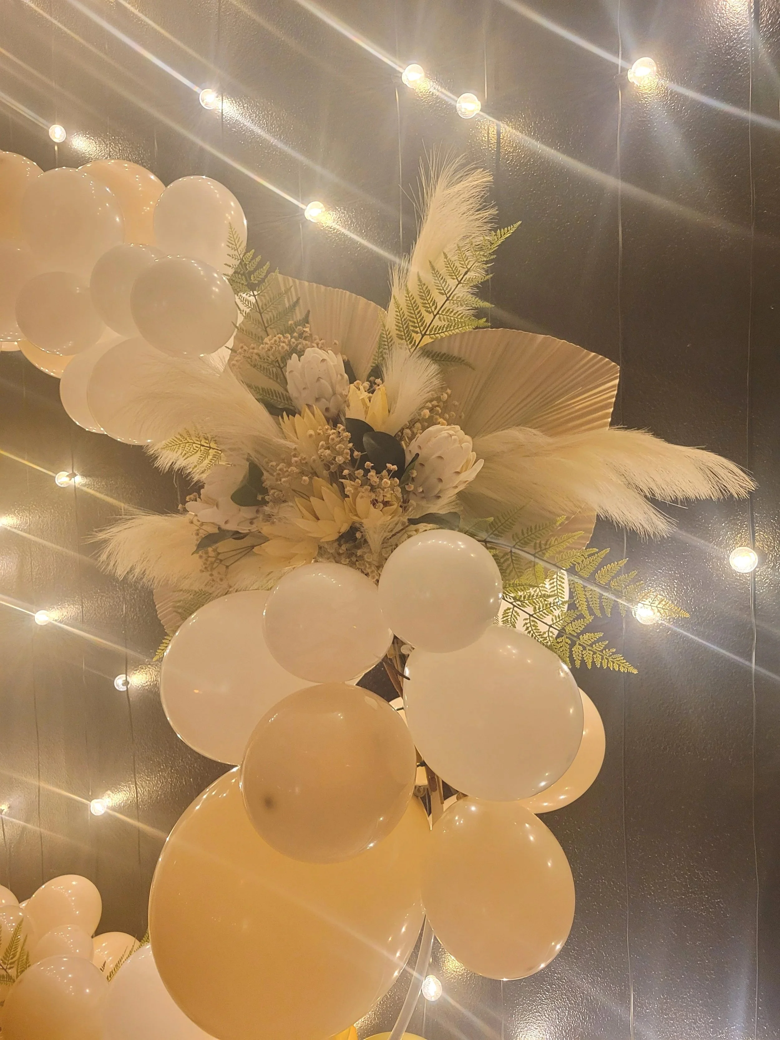 Decorative floral arrangement with white balloons, ferns, and dried leaves on a dark wall illuminated by string lights.