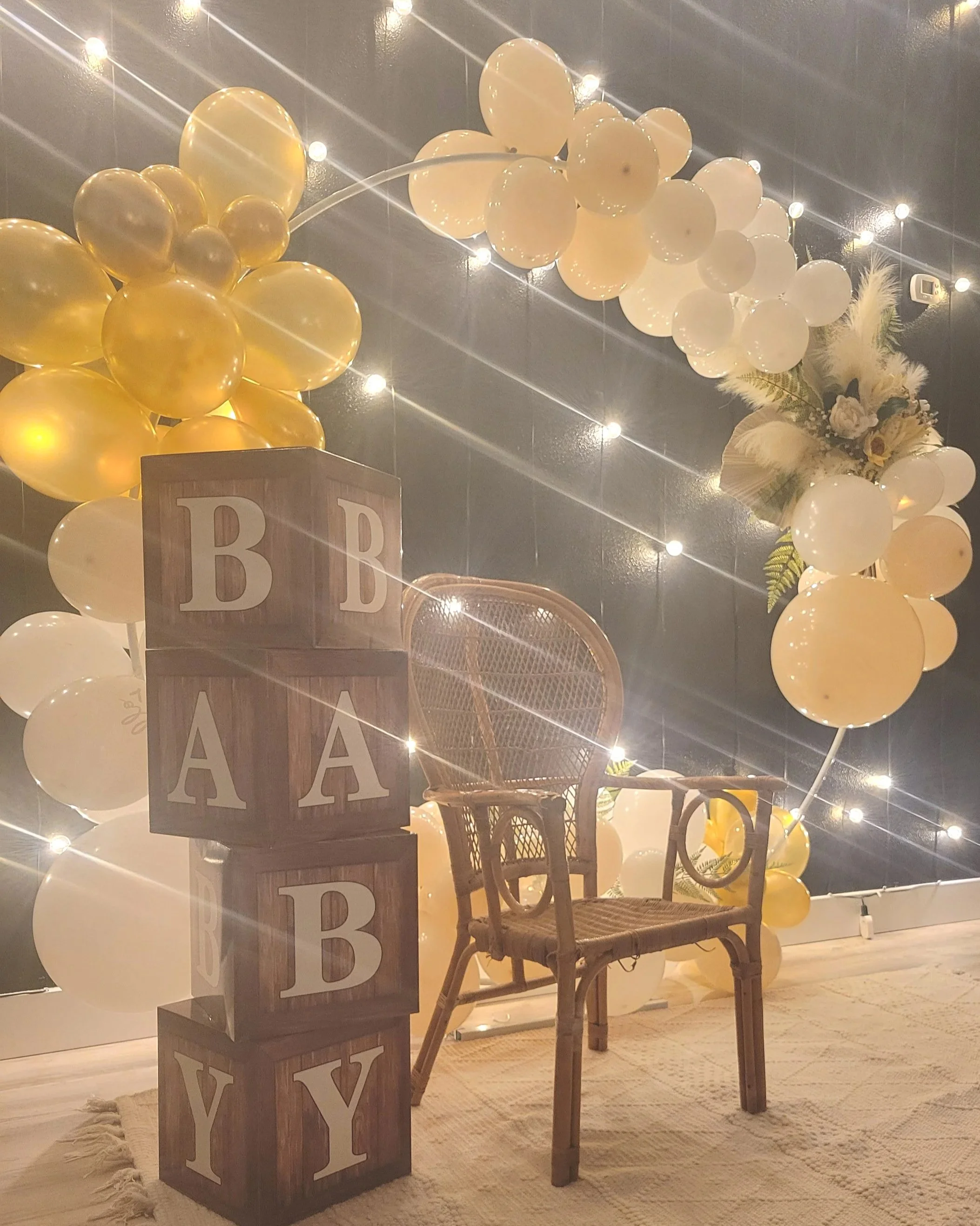 Decorative setup with yellow and white balloons, string lights, wooden blocks spelling "BABY" with letters B, A, B, Y, a wicker chair, and a floral arrangement.