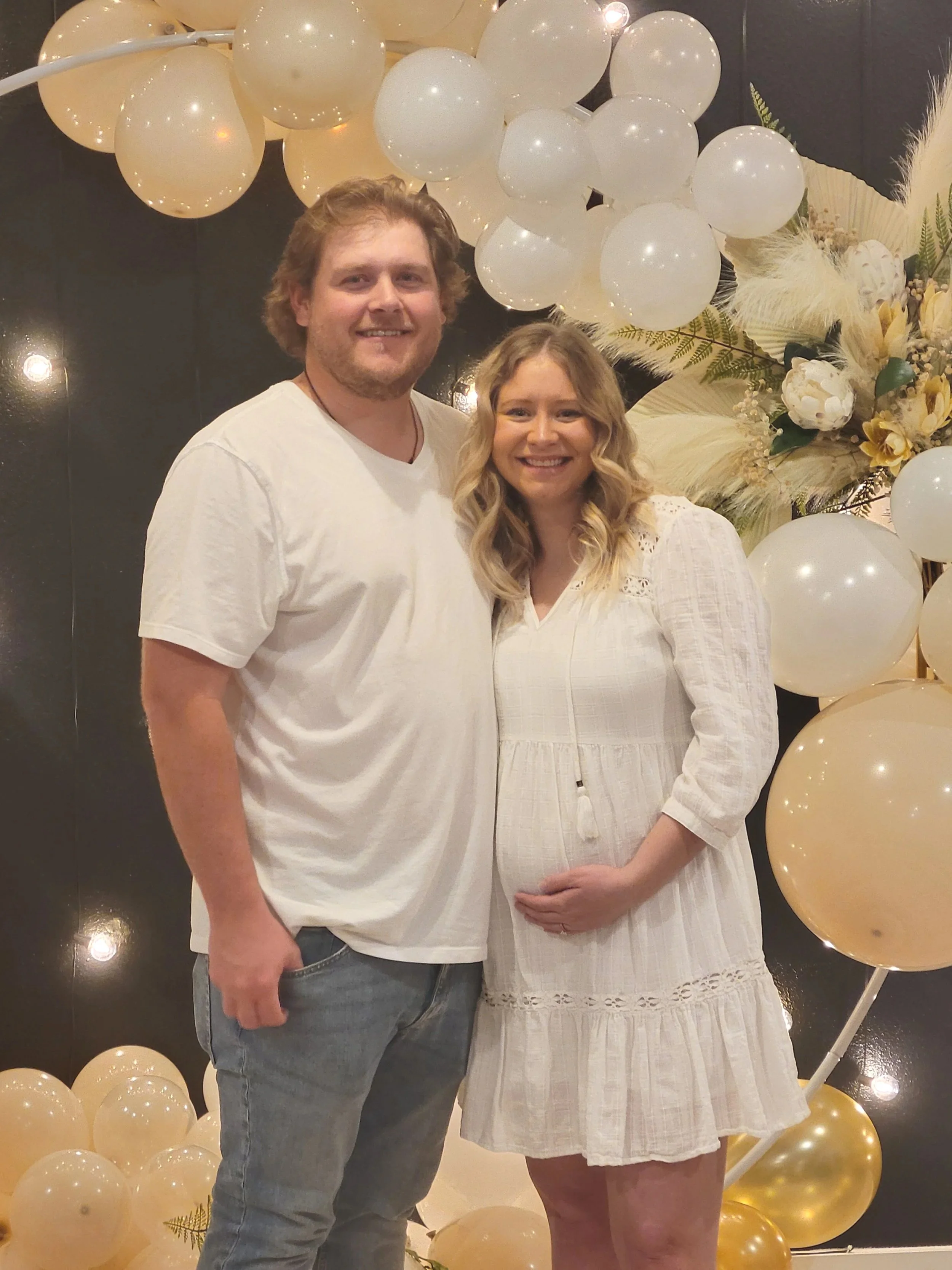 A smiling couple, one pregnant woman and one man, standing in front of a decorative balloon backdrop with white, cream, and gold balloons and white flowers.