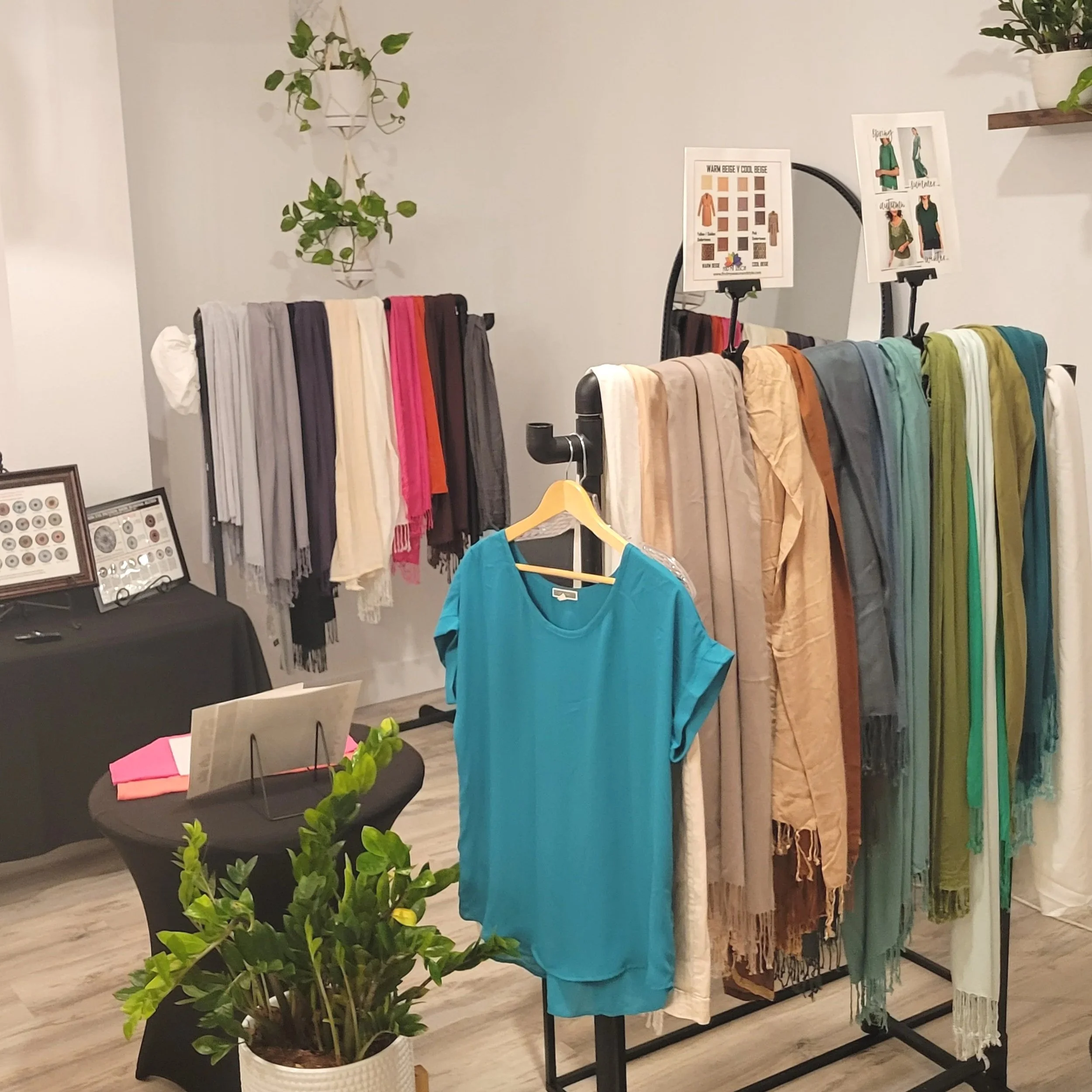 Display of colorful scarves on black racks, with a turquoise shirt hanging in front, inside a shop setting with framed items on a table and decorative plants.
