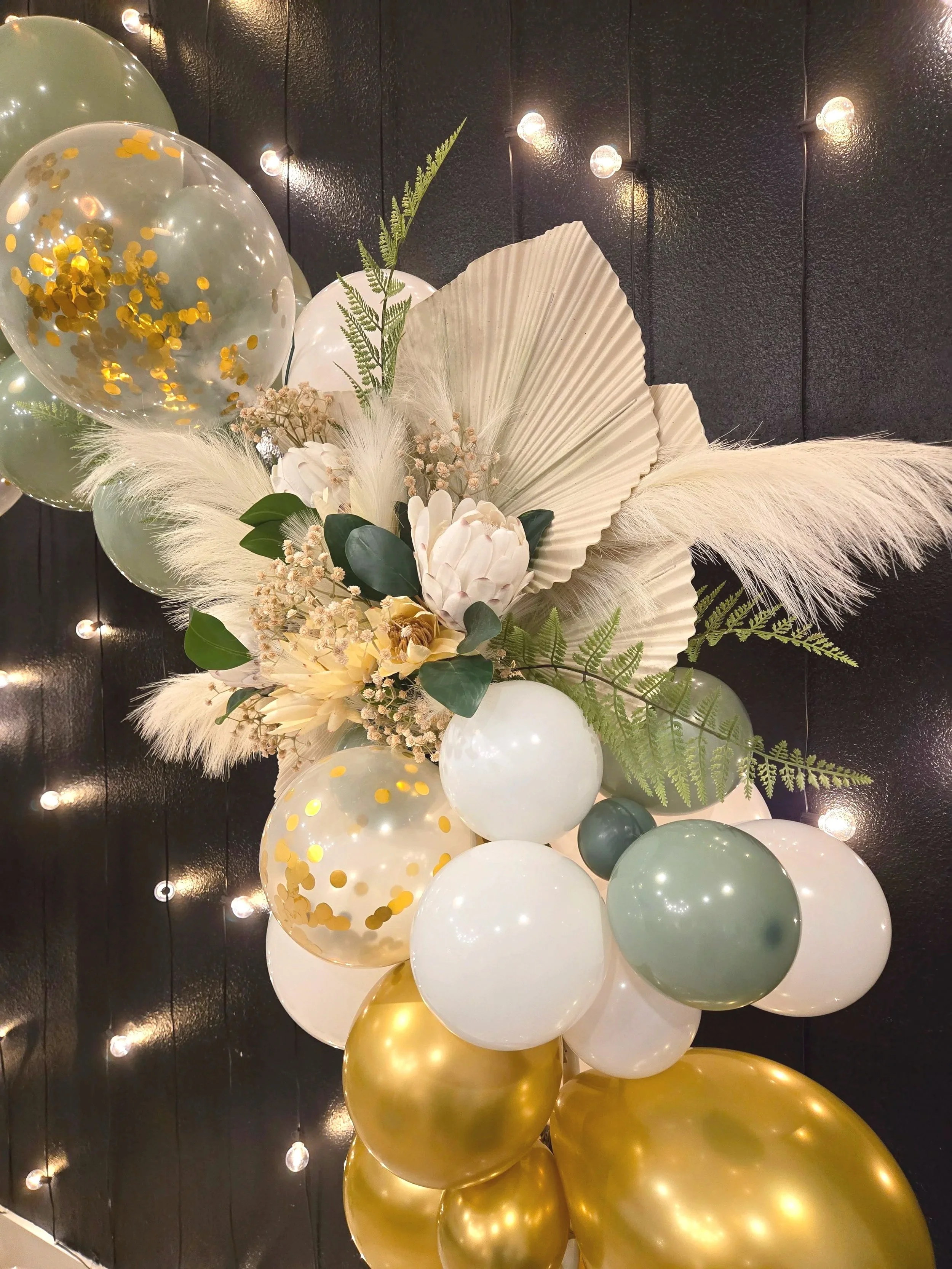 Decorative centerpiece with balloons in gold, white, and green, and a floral arrangement featuring white protea, dried flowers, ferns, and pampas grass, against a dark wall with string lights.