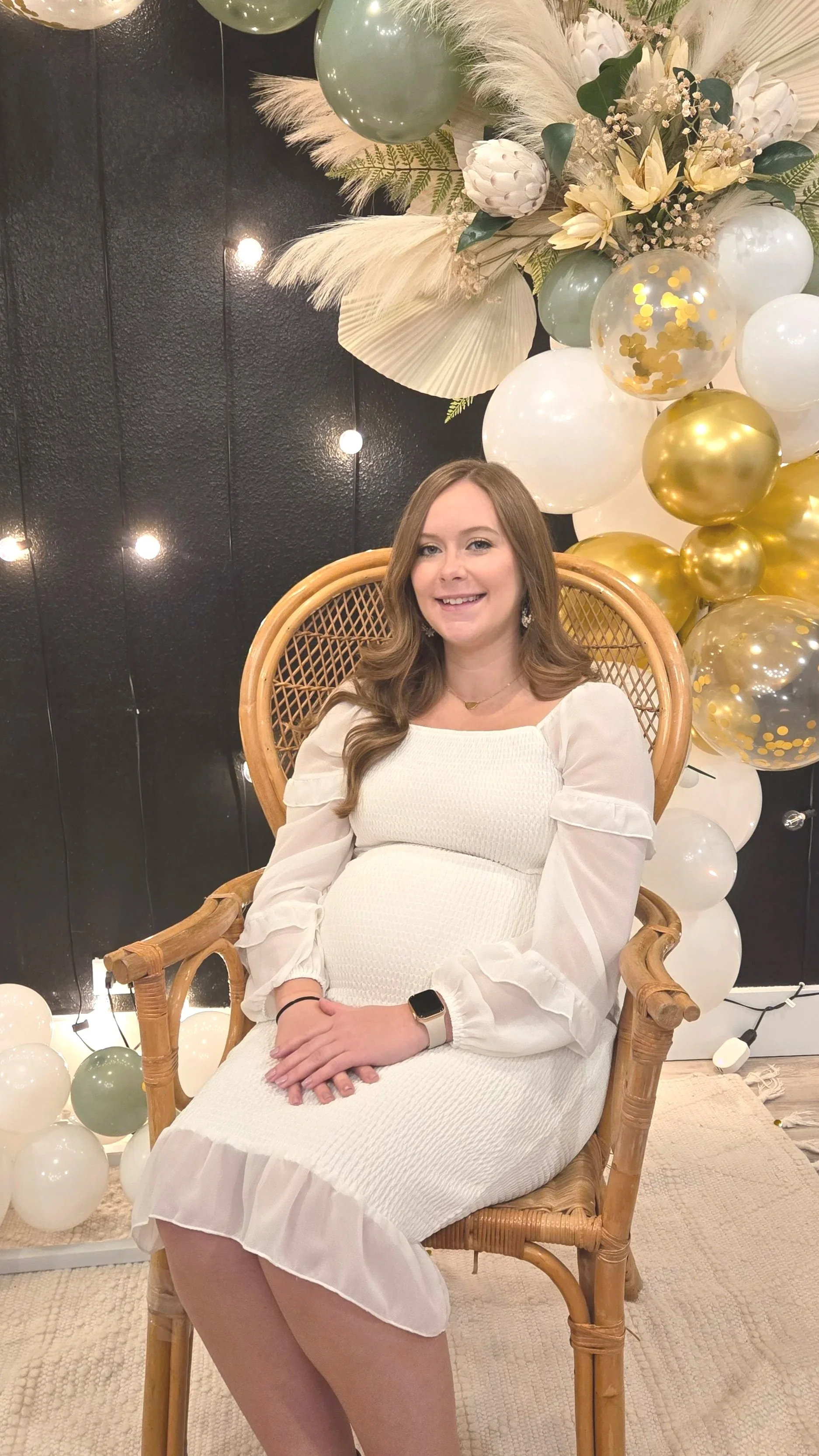 A woman sitting in a woven rattan chair during a celebration, with decorative balloons and floral arrangements in the background.